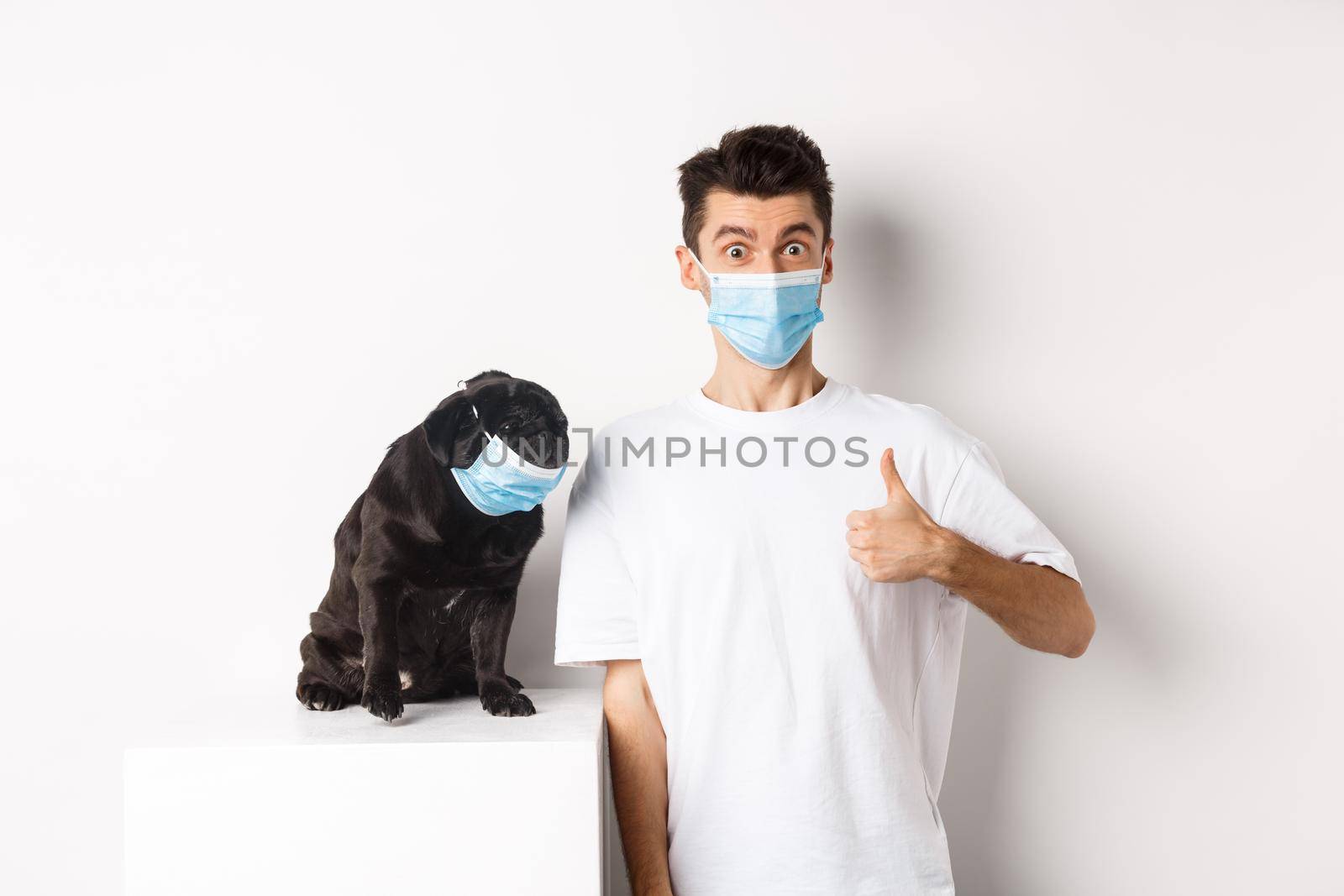 Covid-19, animals and quarantine concept. Image of funny young man and small dog in medical masks, owner showing thumb up in approval, praise something, white background.