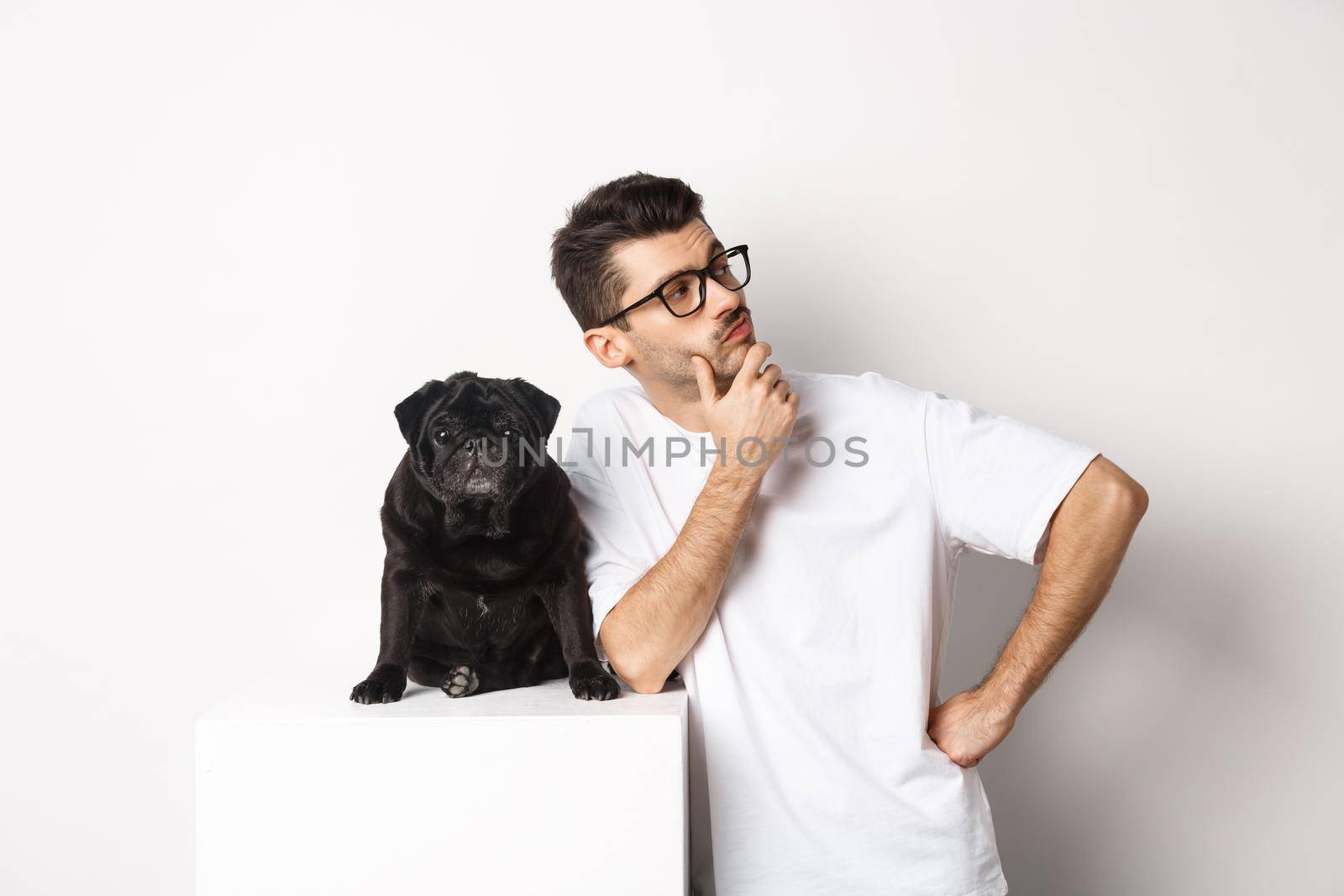 Image of handsome young man, pet owner standing with his dog and looking right at logo, posing with pug against white background.