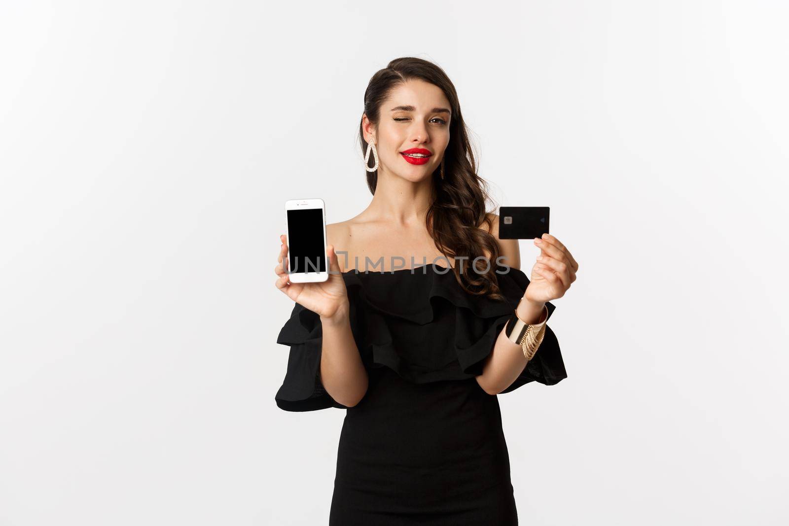 Fashion and shopping concept. Beautiful woman with red lips, winking at camera, showing smartphone screen and credit card, buying online, white background.