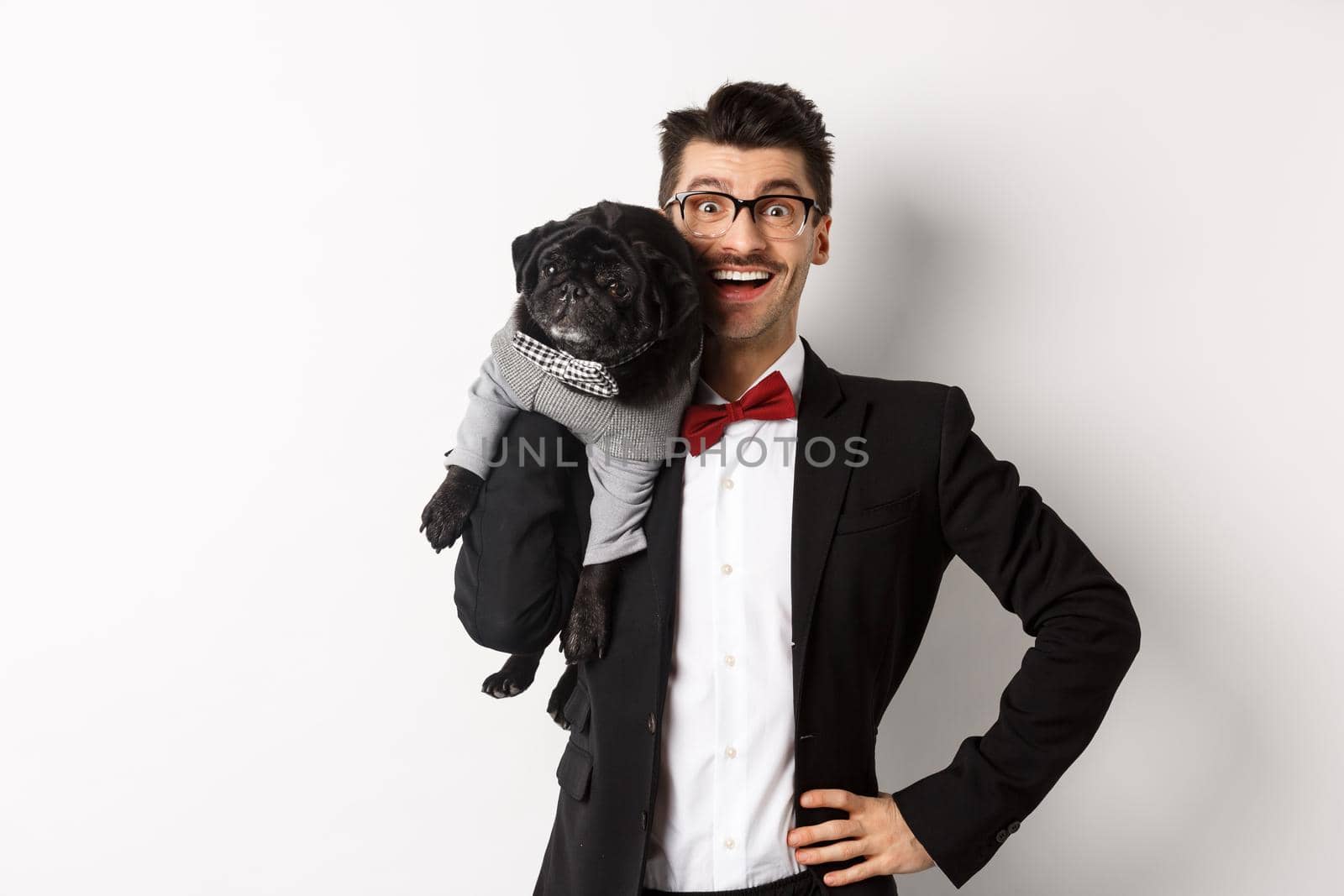 Handsome young man in suit and glasses holding cute black pug dog on shoulder, smiling happy at camera, wearing party outfits, white background.