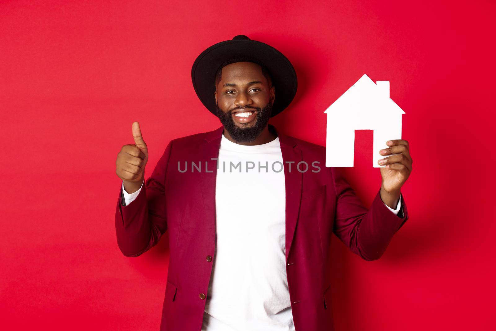 Real estate. Stylish african american bearded man showing home maket, thumb up, recommending agency or apartment complex, standing over red background.