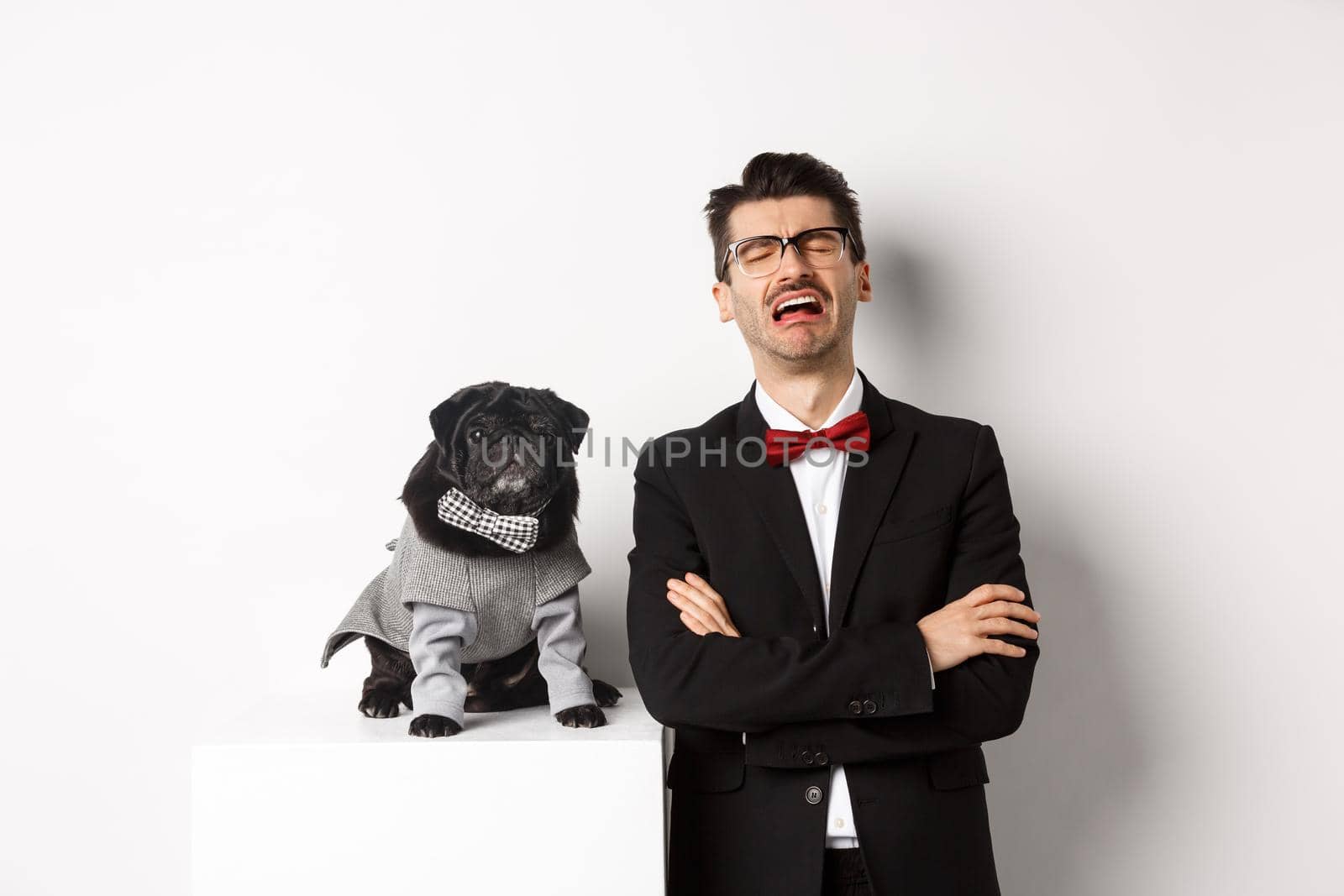 Animals, party and celebration concept. Sad dog owner crying, wearing suit, standing near cute black pug in costume, standing over white background by Benzoix