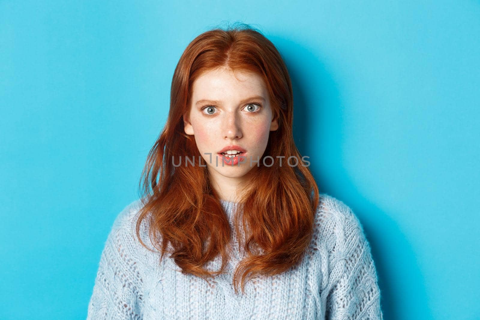 Close-up of amazed redhead girl staring at camera with complete disbelief, standing shocked against blue background.