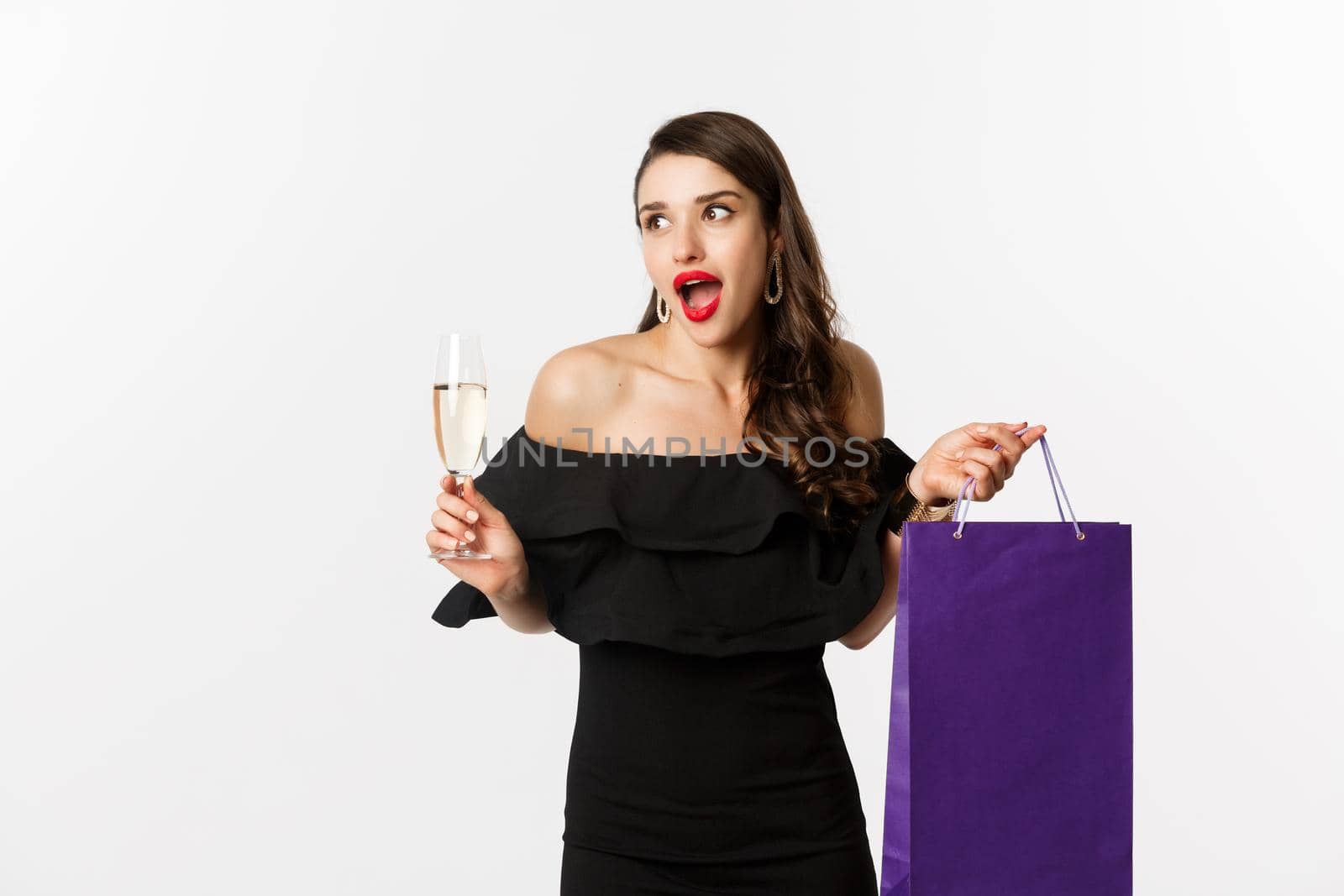 Excited woman shop and drink champagne, holding shopping bag, looking amazed, standing against white background.