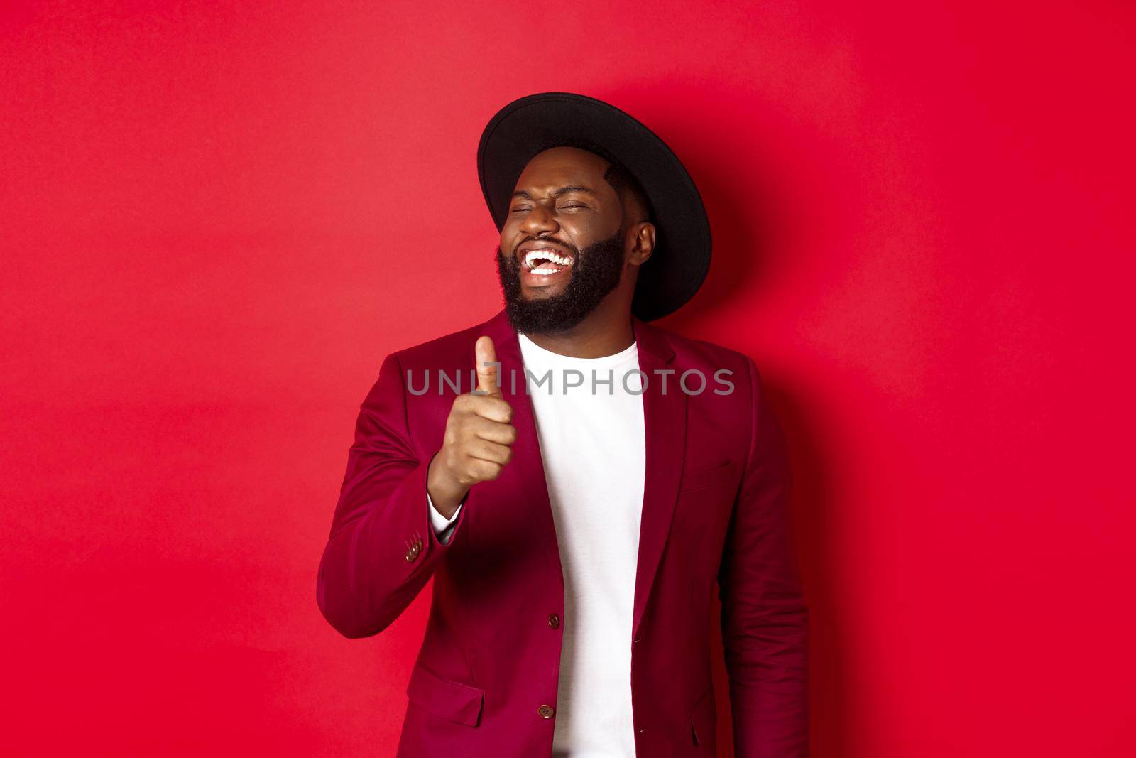Handsome african american man having fun, showing thumb up and laughing from good joke, standing against red background by Benzoix