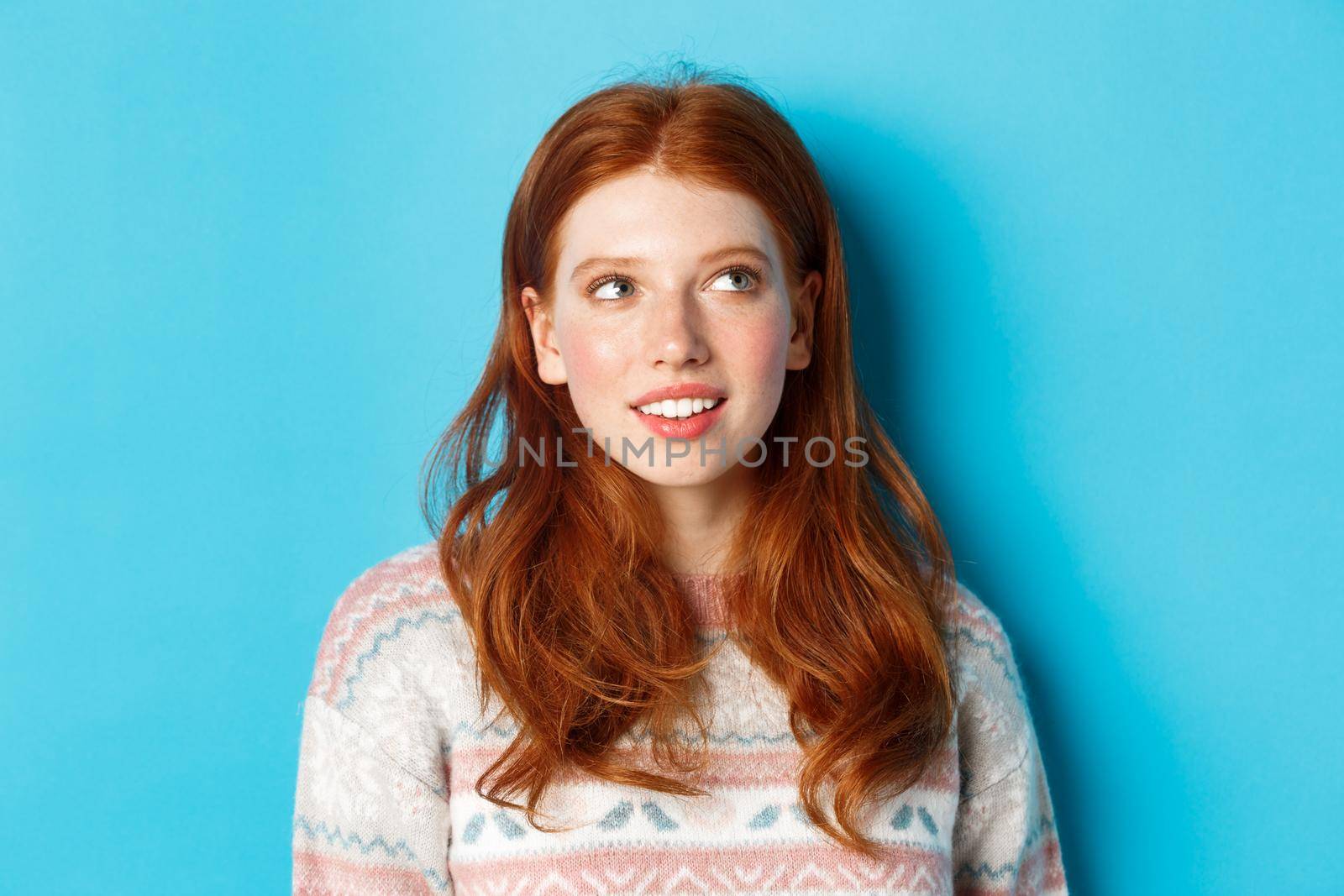 Close-up of dreamy redhead girl imaging something, staring at upper right corner and smiling, standing in winter sweater against blue background.