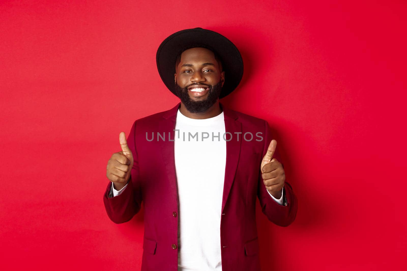 Christmas shopping and people concept. Handsome Black man smiling satisfied, showing thumbs-up, like and agree, approve something, standing against red background.