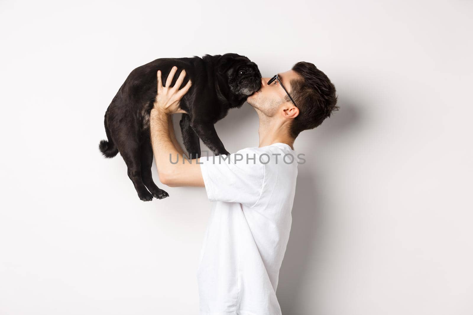 Profile of handsome young man kissing small cute dog face. Hipster guy loving his pug, standing over white background by Benzoix