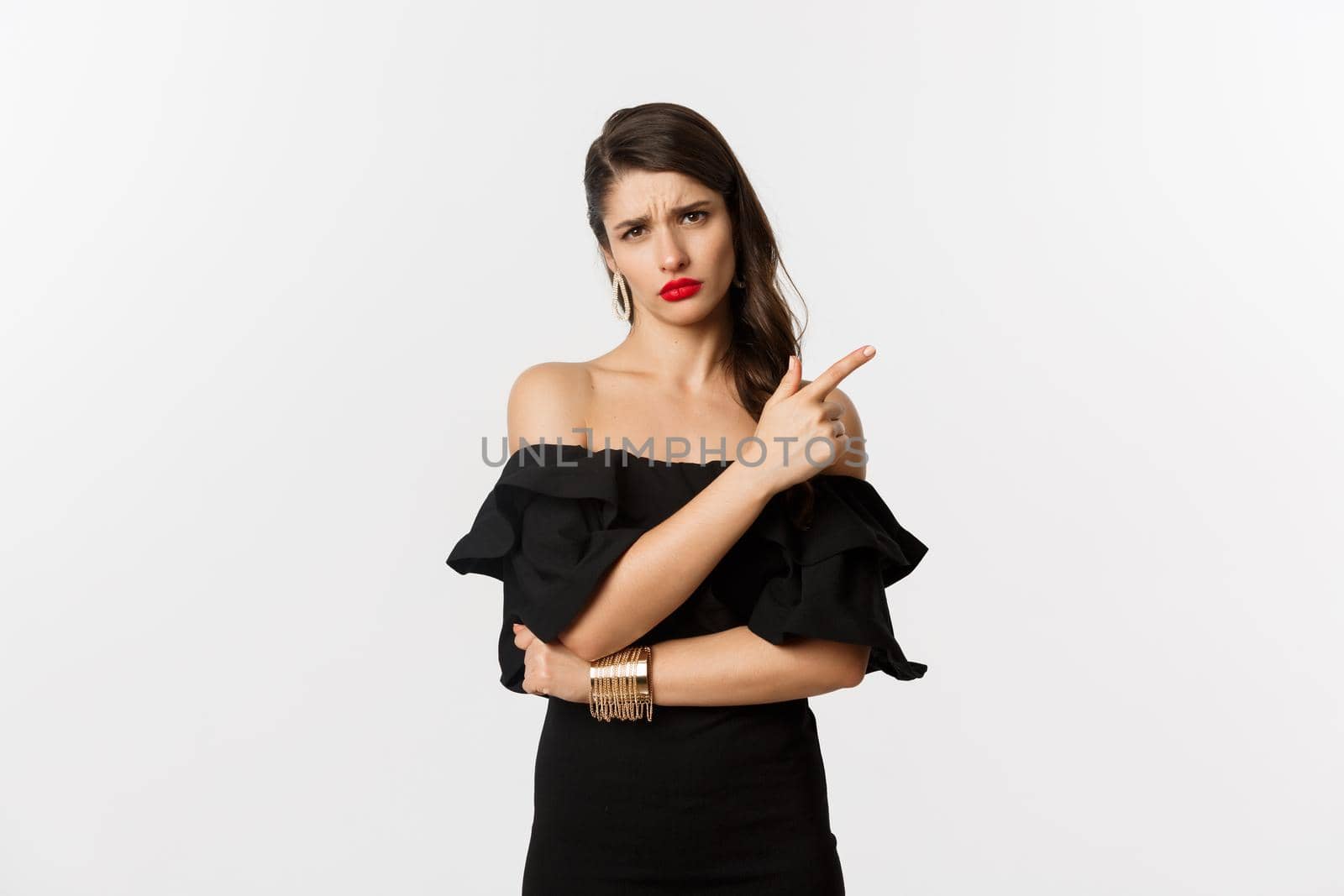 Fashion and beauty. Skeptical glamour woman with red lips, black dress, pointing finger right at something lame and boring, standing over white background.
