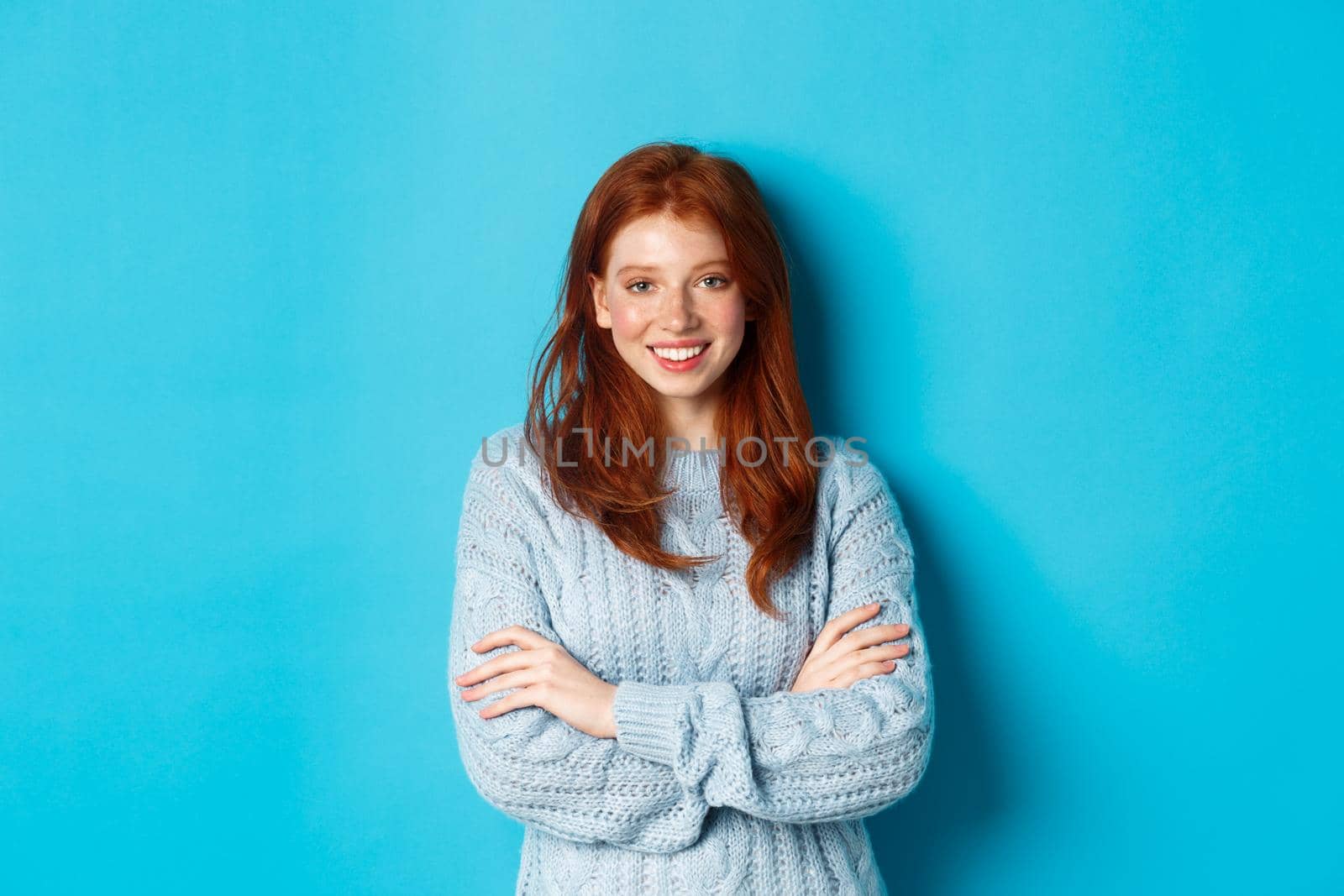 Attractive redhead girl in sweater smiling and staring at camera, standing confident against blue background.