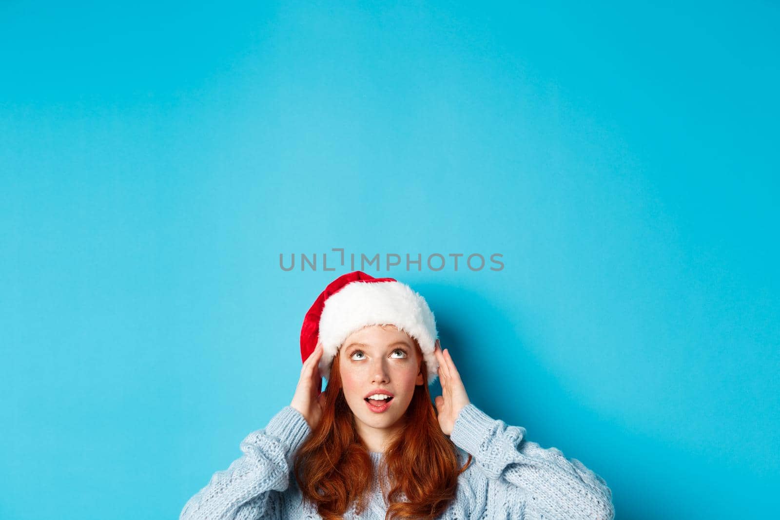 Winter holidays and Christmas eve concept. Head of cute redhead girl in santa hat, appear from bottom and looking up at copy space, staring logo, standing over blue background.