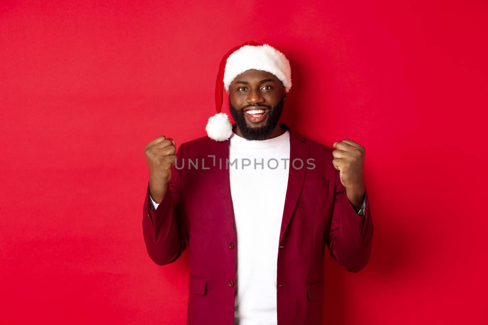 Christmas, party and holidays concept. Excited Black man celebrating New Year, wearing santa hat, clench fists satisfied, winning and rejoicing, standing against red background by Benzoix