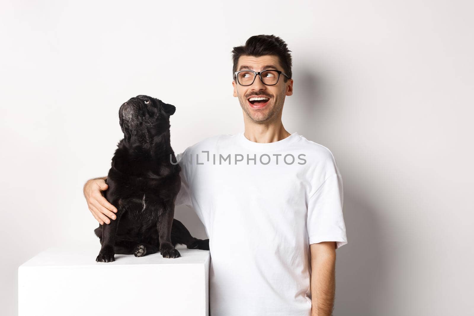 Amazed young man in glasses hugging his dog, pet owner and pug staring at upper left corner promo offer, standing over white background.