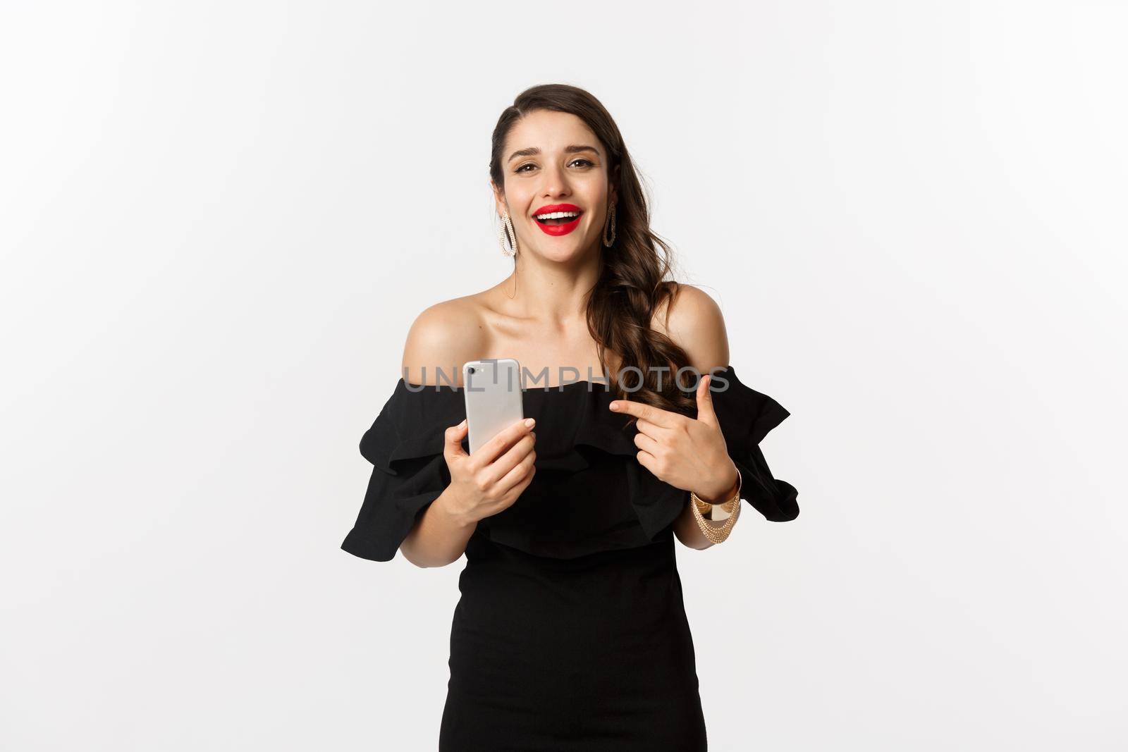 Online shopping concept. Satisfied pretty woman in black dress, smiling pleased and pointing at mobile phone, standing over white background.
