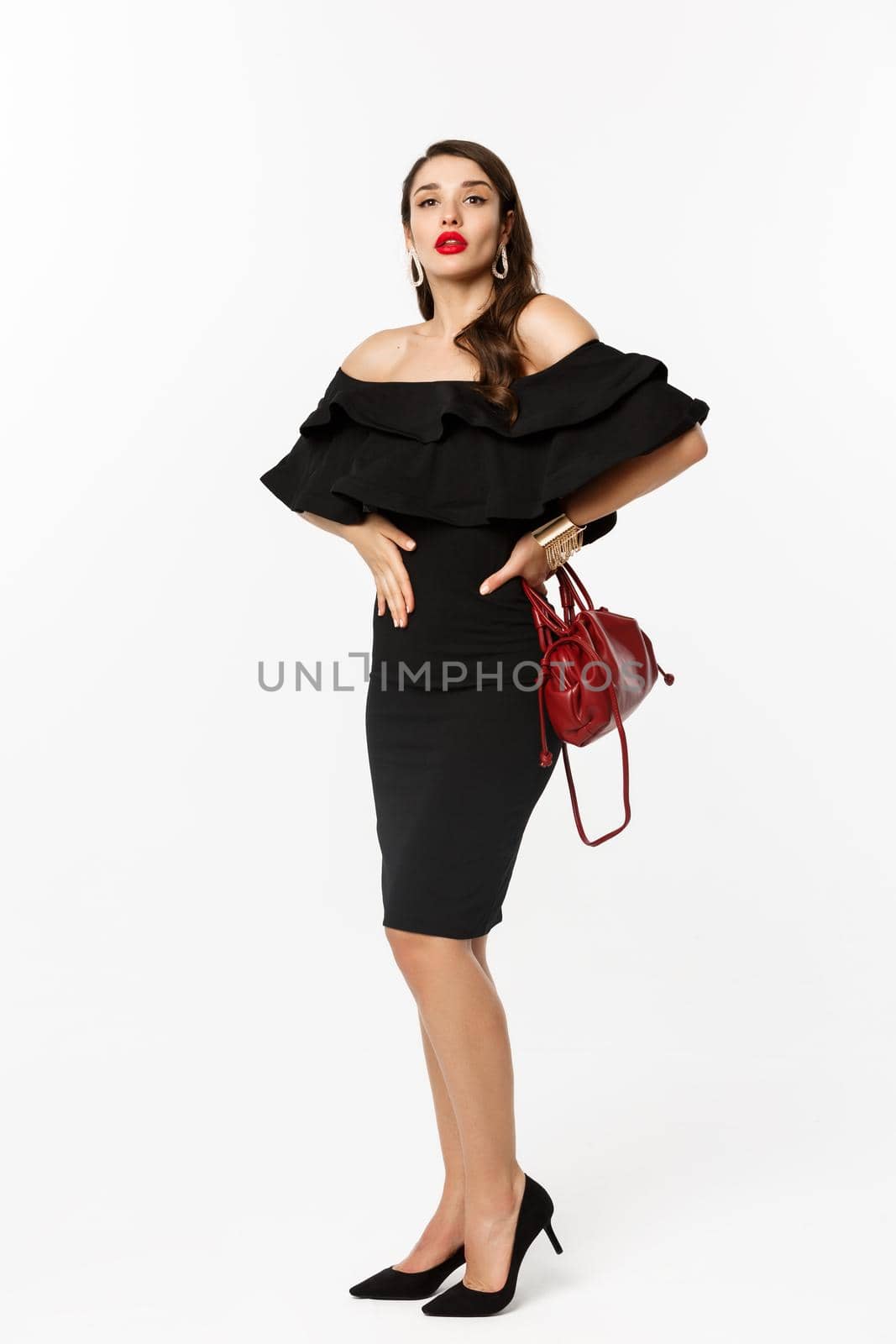 Beauty and fashion concept. Full length of elegant young woman going on party in black dress, high heels, looking confident and sassy at camera, white background.