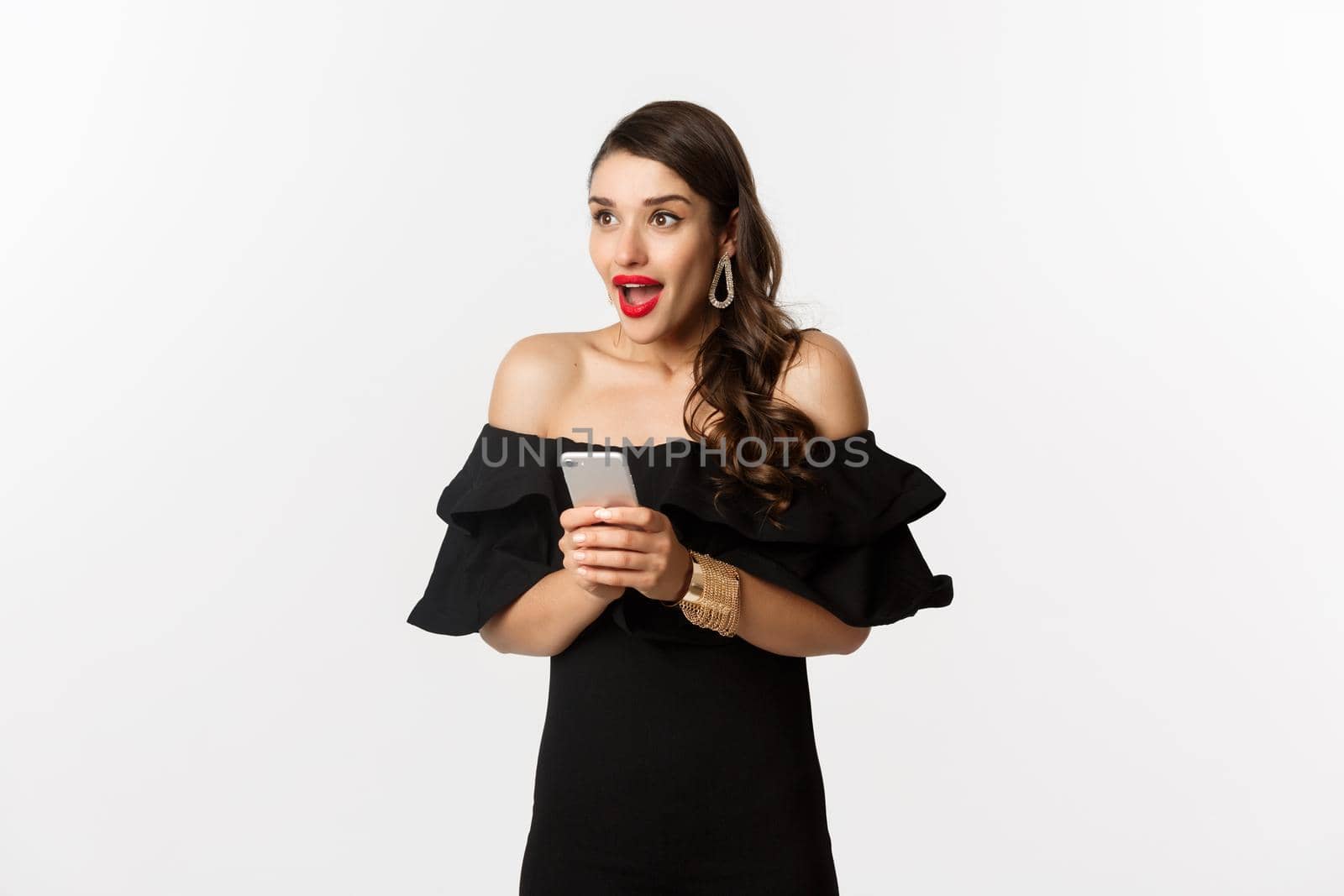 Image of excited woman in black glamour dress, red lipstick, using mobile phone and looking left amazed, standing over white background.