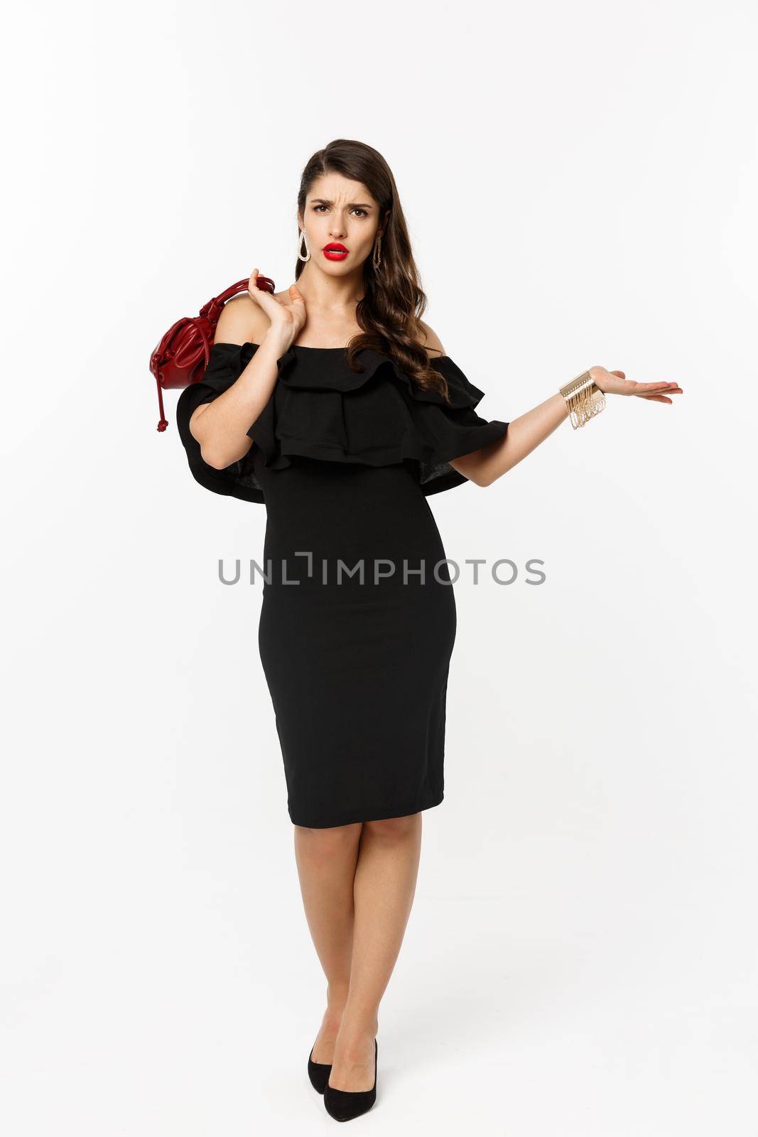 Beauty and fashion concept. Full length of arrogant and sassy woman in black dress, holding purse on shoulder and stroll forward at camera, posing over white background.