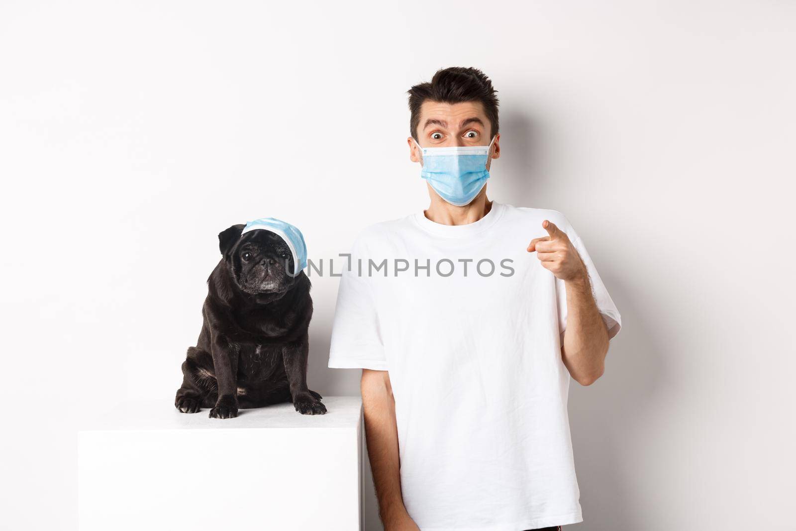 Covid-19, animals and quarantine concept. Happy dog owner and cute pug wearing medical masks, man pointing finger at camera amazed, white background.