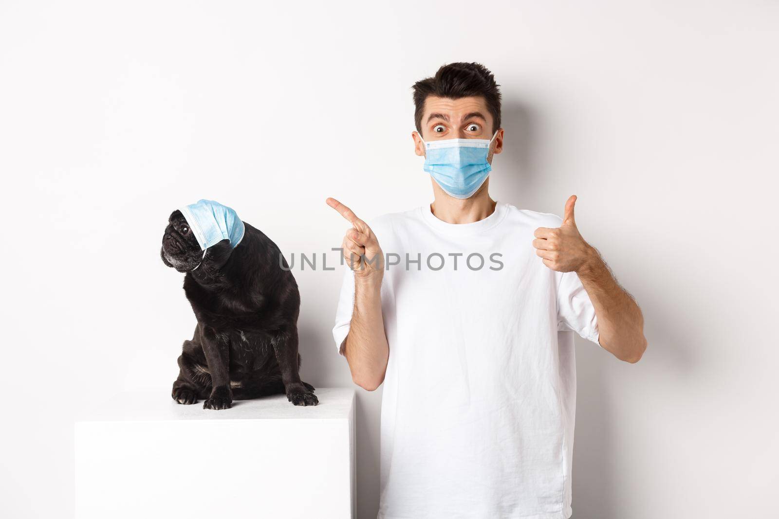 Covid-19, animals and quarantine concept. Young man and black dog wearing medical masks, pug looking at upper left corner and owner showing thumb-up to praise promo by Benzoix