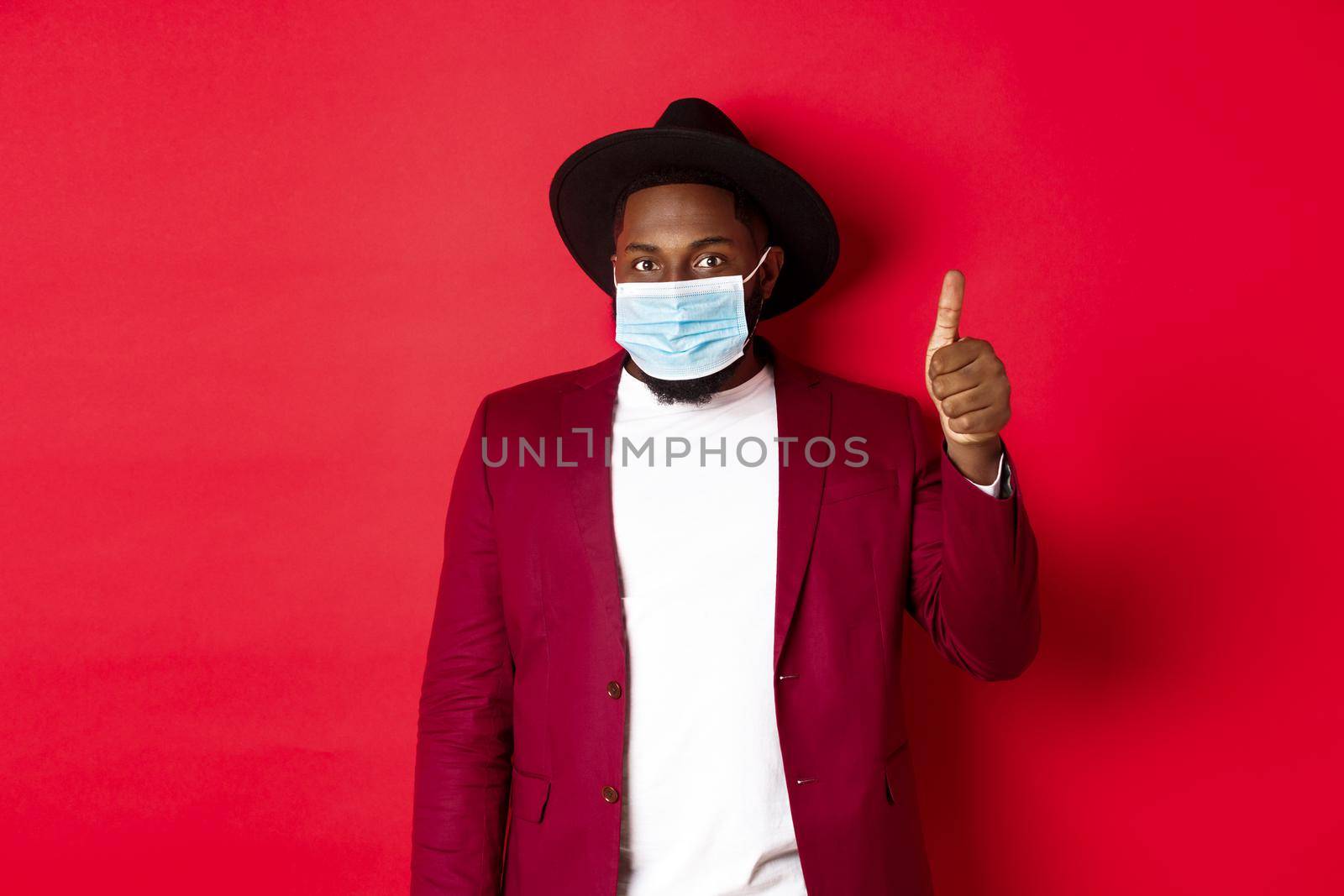 Covid-19 and fashion concept. Stylish african american man in hat and blazer, wearing face mask and showing thumb up, standing over red background.