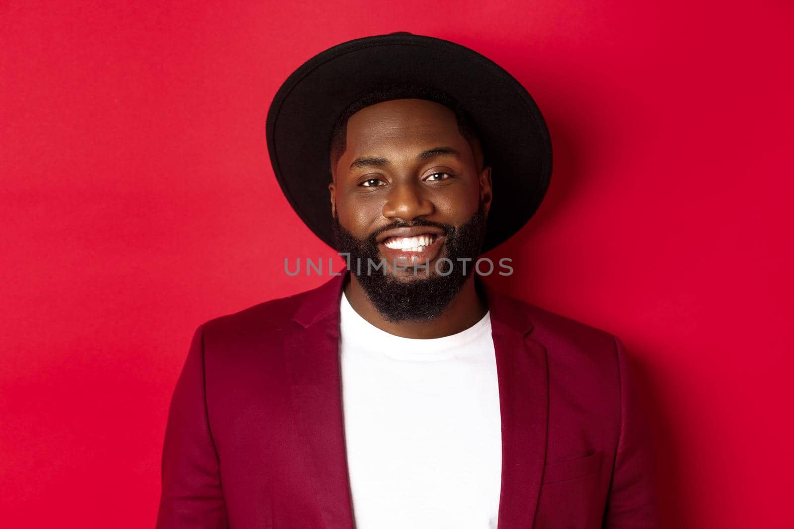 Close-up of handsome african american man with beard, wearing party blazer and stylish hat, smiling at camera, red background.