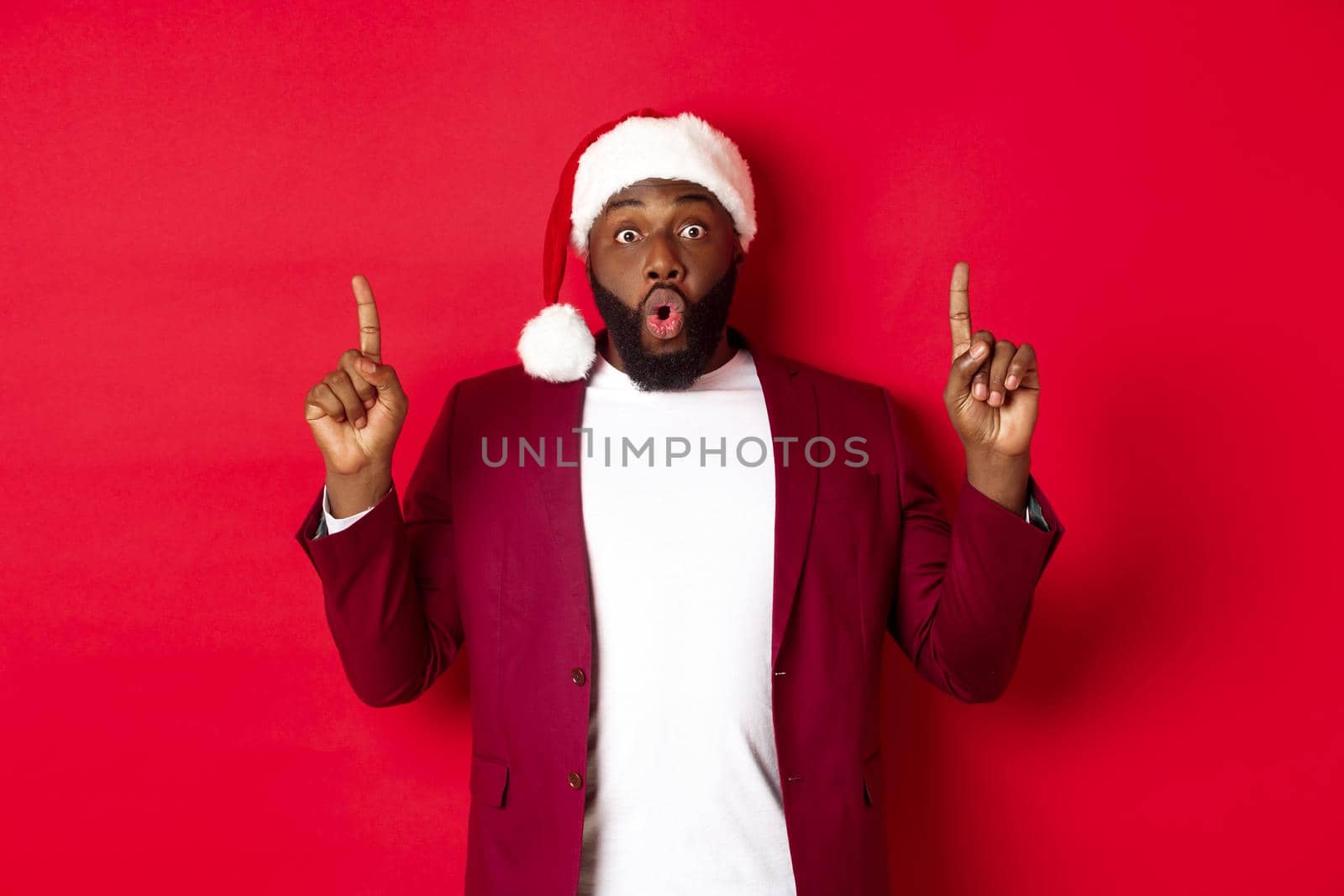 Christmas, party and holidays concept. Impressed Black man showing offer, pointing fingers up at logo, saying wow, standing against red background by Benzoix