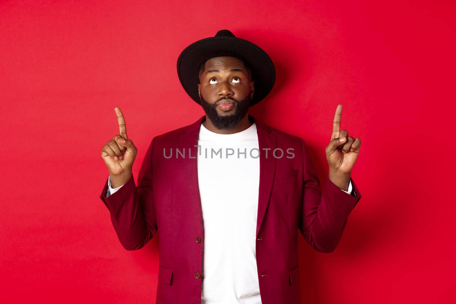 Winter holidays and shopping concept. Curious african american guy in party clothing looking and pointing fingers up, reading promo sign, standing over red background.