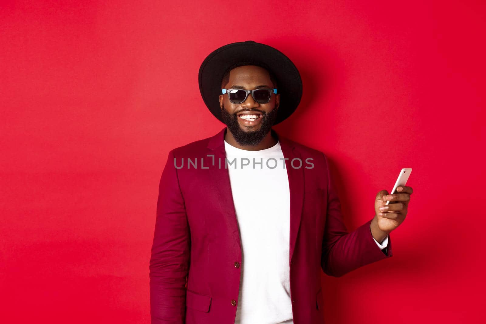 Handsome african american man shopping online, making an order on smartphone and smiling, standing over red background.