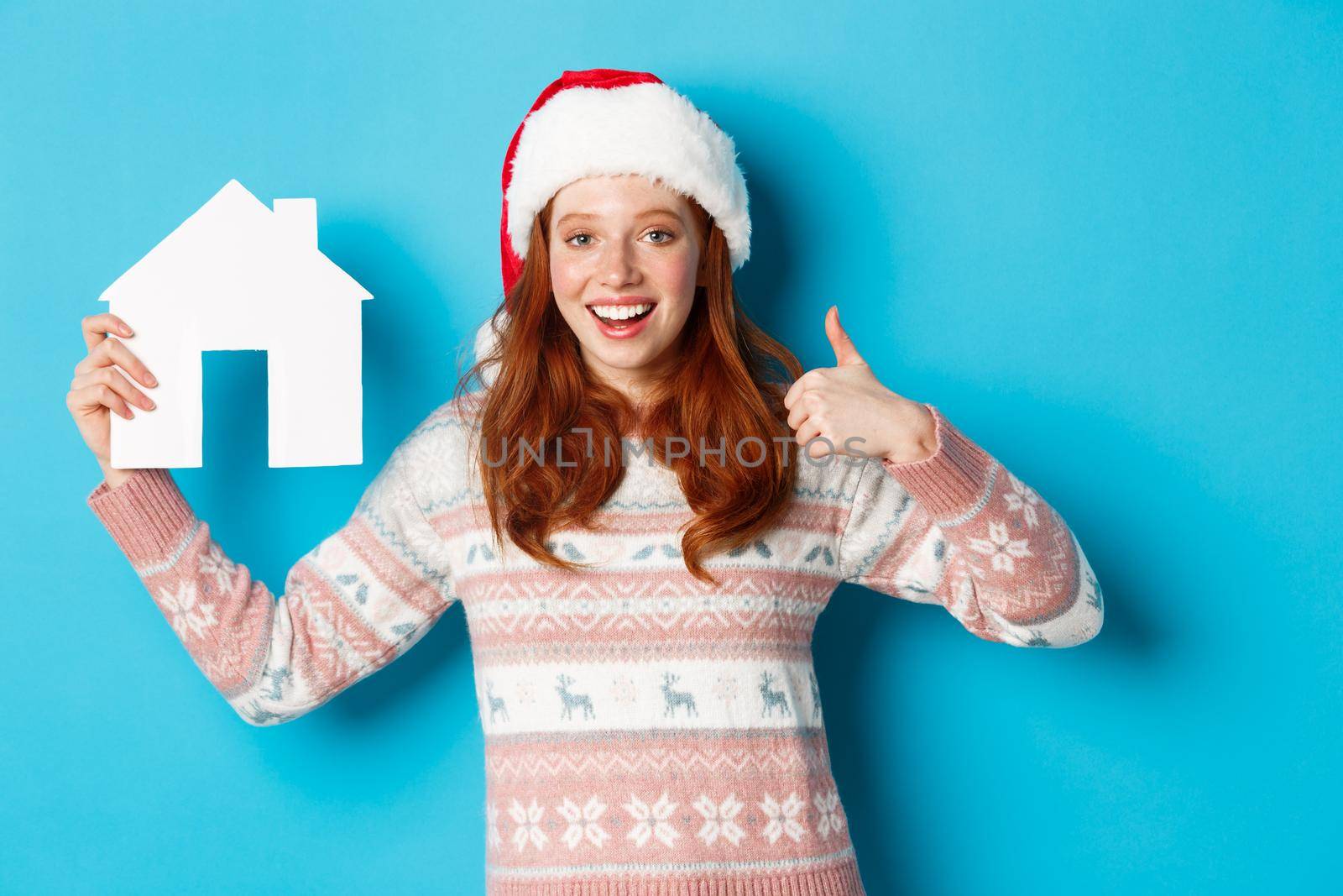 Holiday promos and real estate concept. Satisfied female model with red wavy hair, wearing santa hat and sweater, showing paper house model and thumbs-up, blue background.