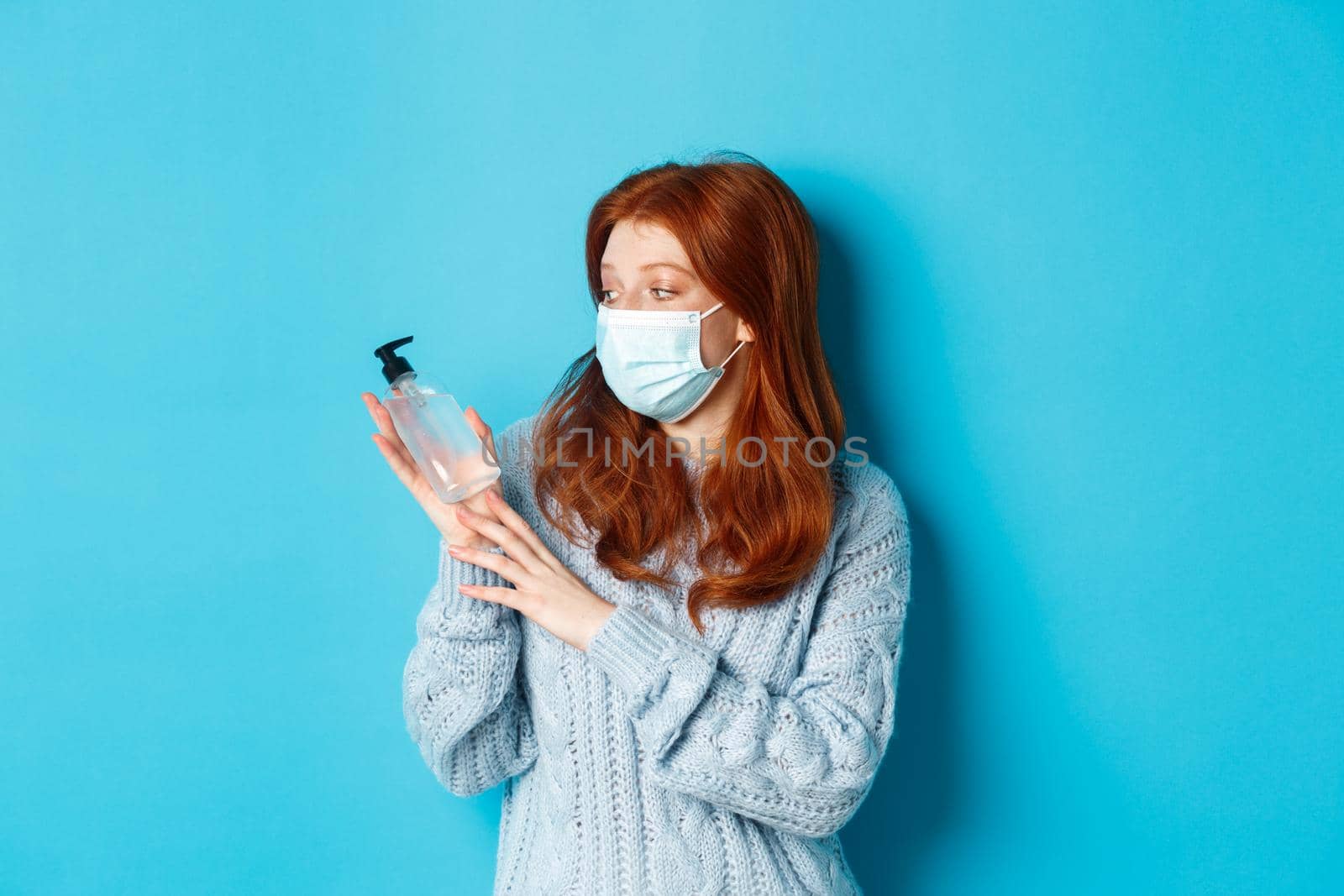 Winter, covid-19 and social distancing concept. Young redhead girl in face mask showing hand sanitizer, demonstrating antiseptic for disinfection, standing over blue background.