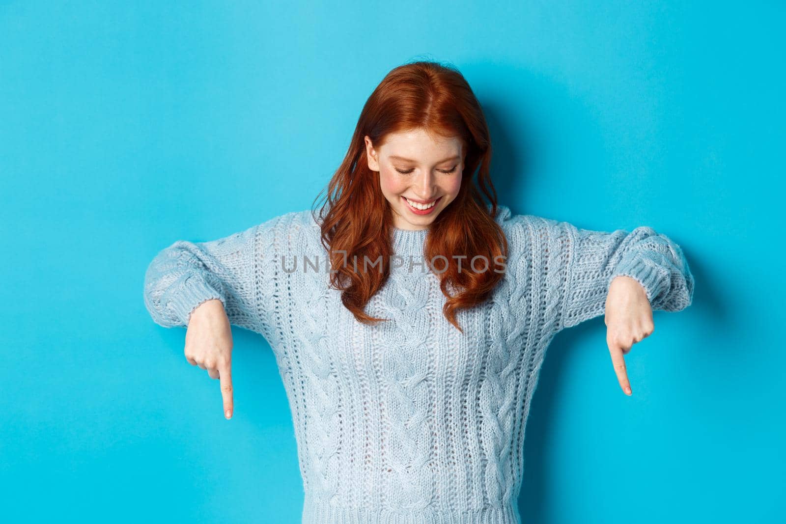 Winter holidays and people concept. Cheerful redhead girl in sweater, pointing fingers down and looking happy at logo, standing over blue background.