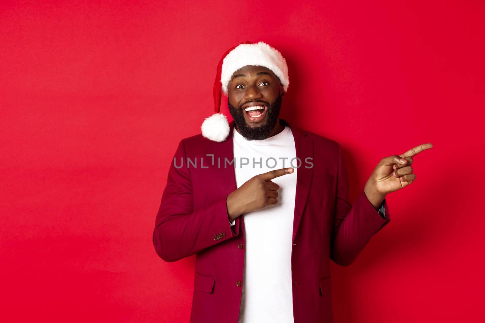 Christmas, party and holidays concept. Cheerful Black man smiling, pointing fingers right and showing advertisement, standing over red background by Benzoix