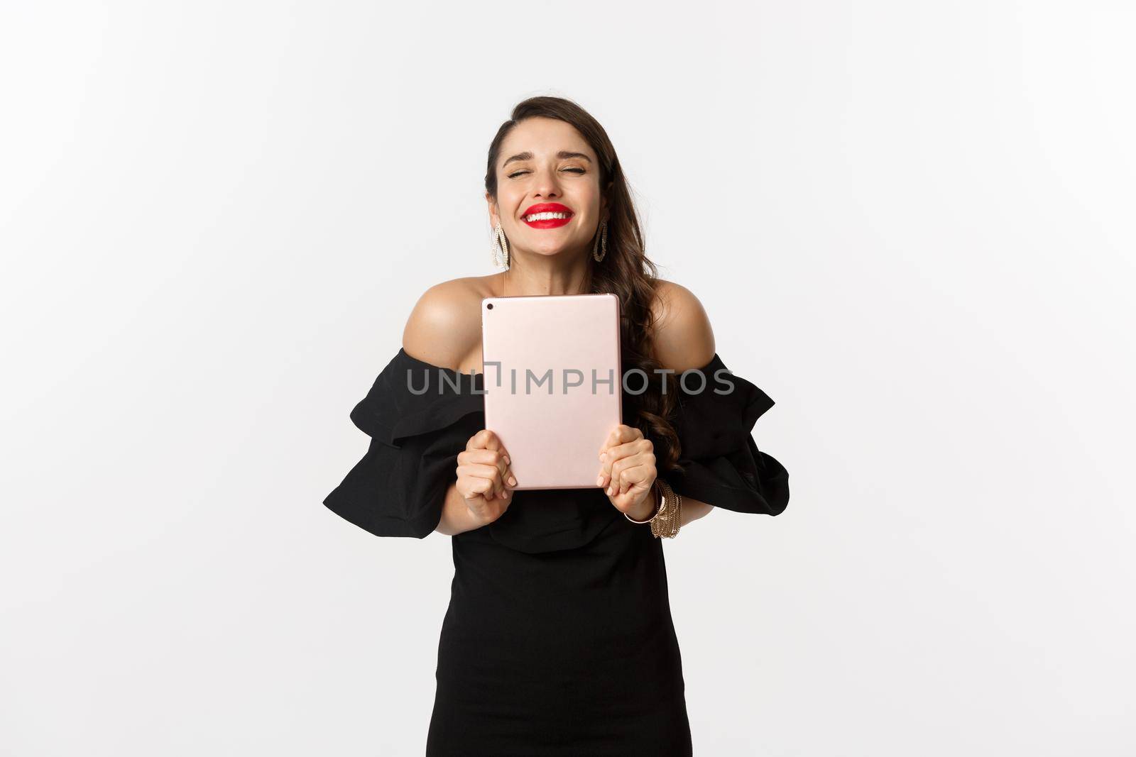Fashion and shopping concept. Happy young woman with red lips, wearing black dress, rejoicing and holding digital tablet, winning prize, white background by Benzoix