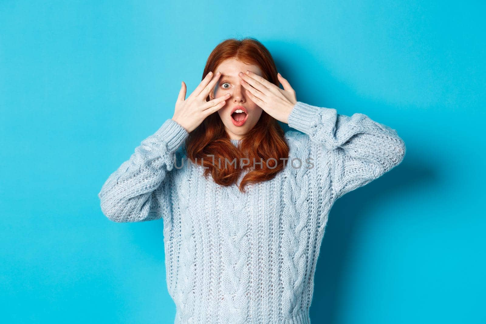 Excited teenage redhead girl open eyes to see holiday surprise, receiving presents, looking amazed at camera, standing over blue background.