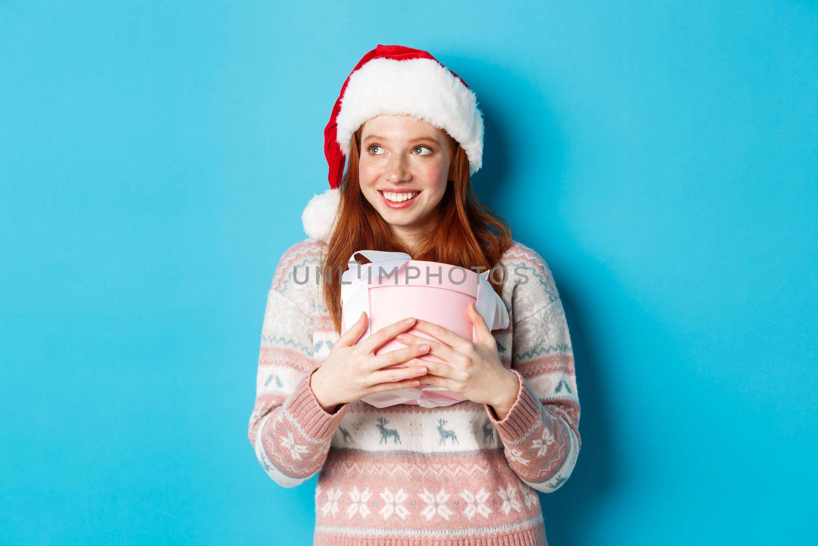 Winter and celebration concept. Dreamy redhead girl in santa hat hugging her christmas gift and looking left, smiling happy, standing over blue background.