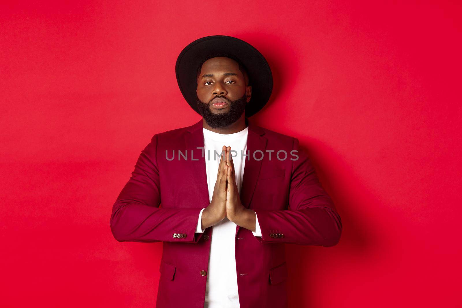 Serious african american man showing respect, holding hands together in namaste or thank you gesture, standing against red background by Benzoix