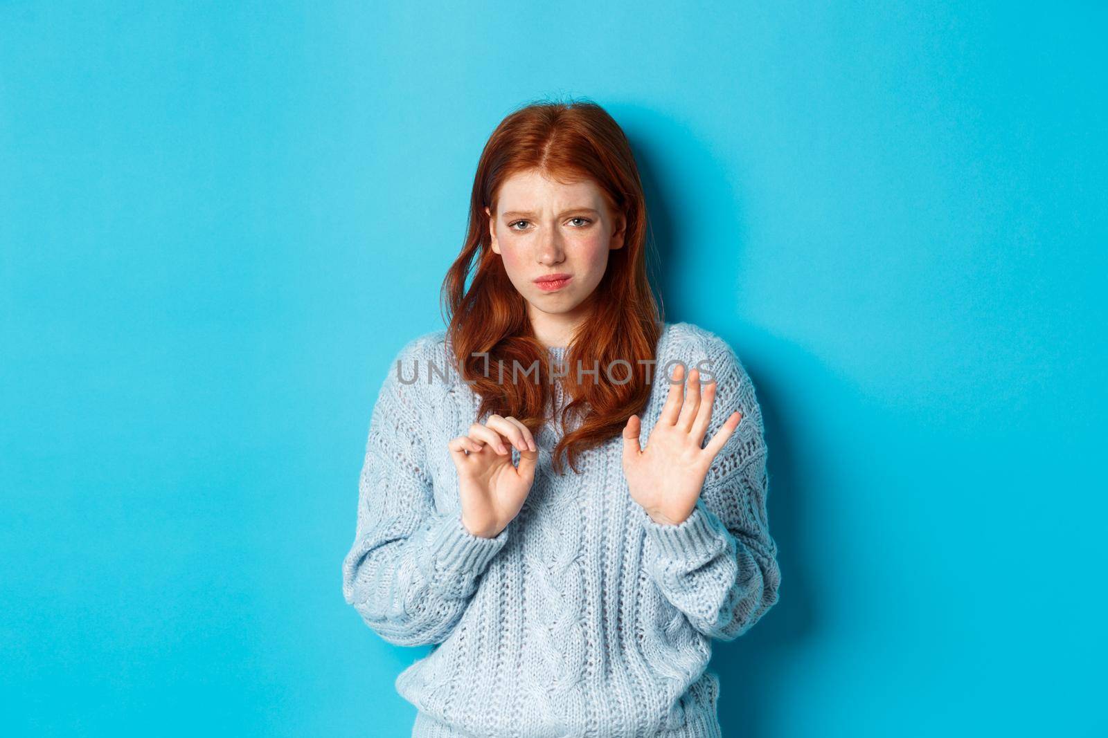 Reluctant redhead girl asking to stay away, shaking hand in rejection gesture, decline offer and grimacing displeased, standing over blue background.