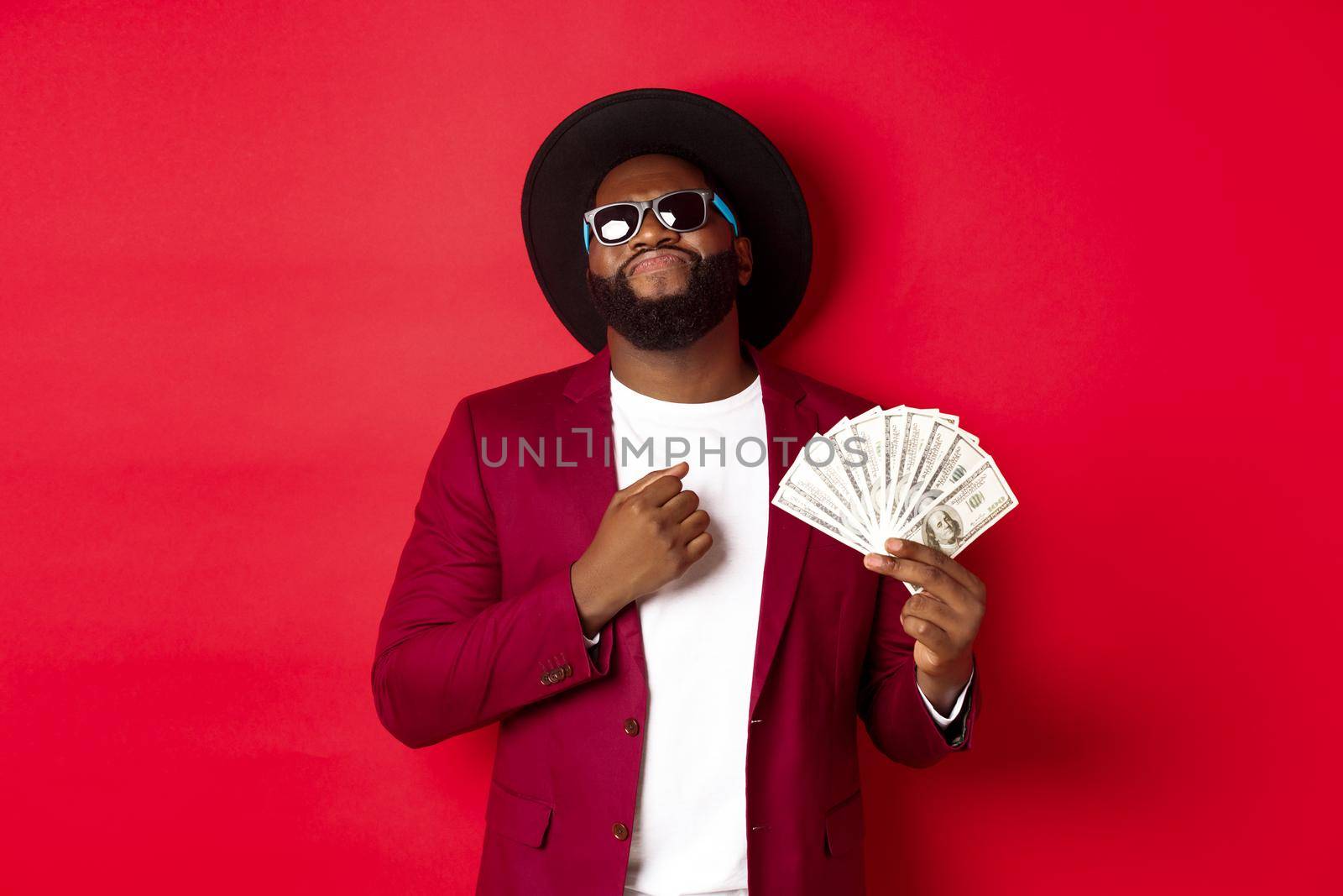 Sassy and cool african american man in sunglasses and hat, pointing at himself and showing dollars, looking confident against red background.