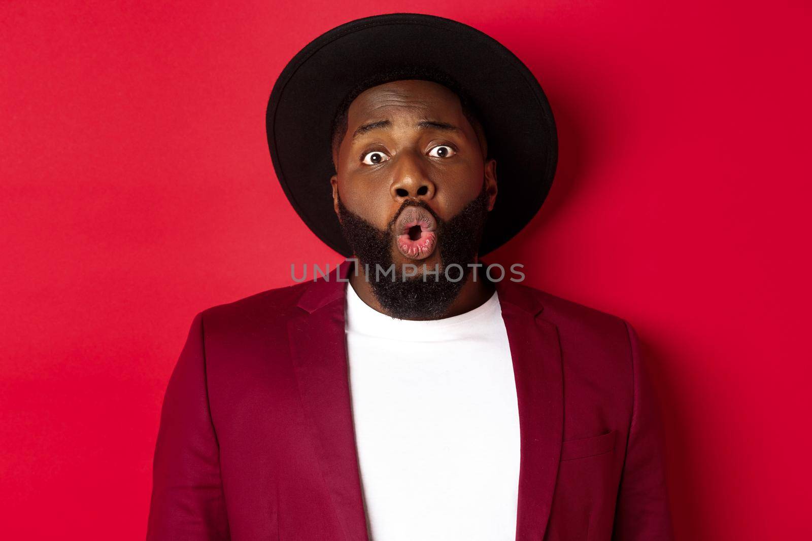 Close-up of surprised Black man staring at camera with wonder, standing against red background by Benzoix