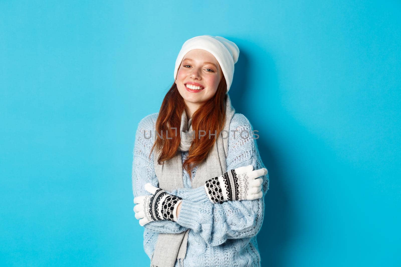 Winter and holidays concept. Smiling redhead girl in beanie, gloves and sweater getting warm after going outside, standing over blue background by Benzoix