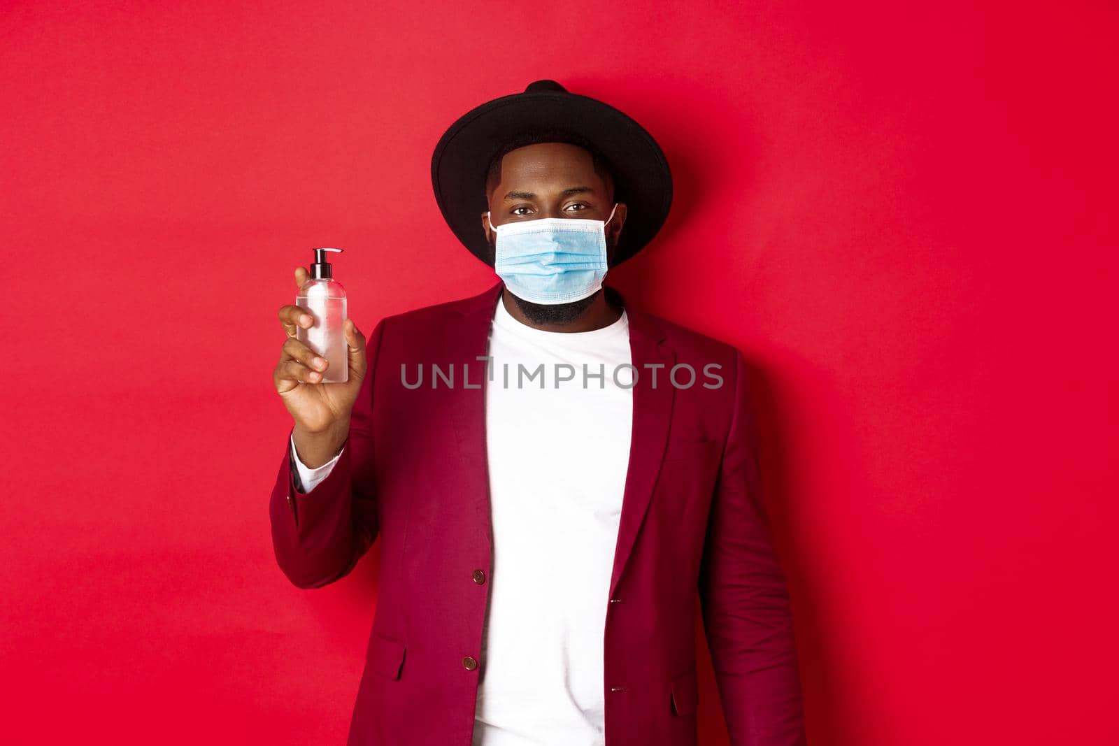 Covid-19, quarantine and holidays concept. Young african american man in medical mask and stylish outfit showing hand sanitizer, recommending to use antiseptic.