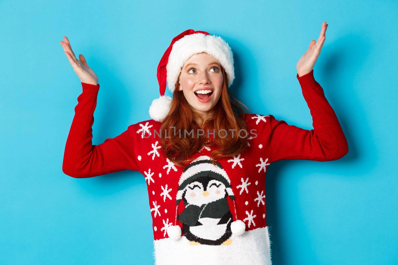 Happy holidays and Christmas concept. Excited redhead girl rejoicing of something falling on her, raising hands up delighted and smiling, wearing Santa hat with xmas sweater, blue background.