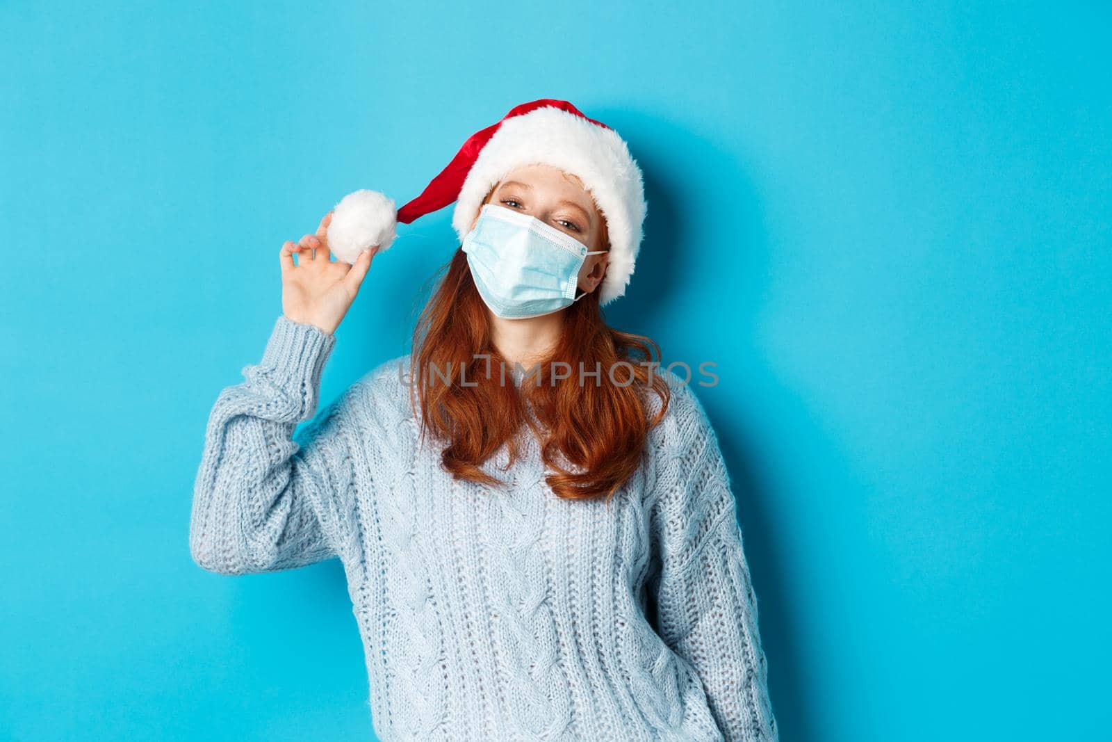 Christmas, quarantine and covid-19 concept. Cheerful teen redhead girl in santa hat and face mask, staring at camera pleased, standing confident against blue background.