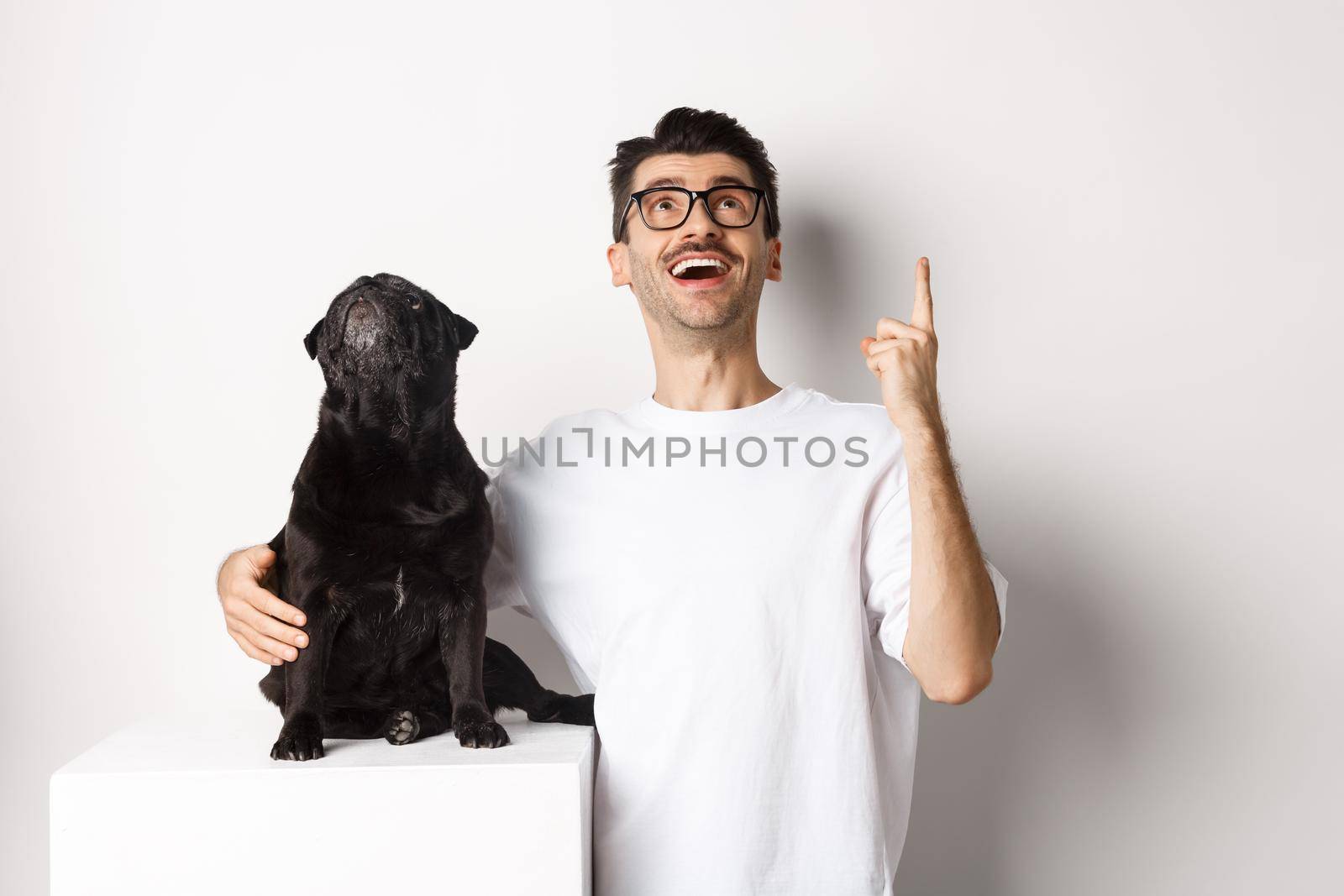 Image of happy young man and dog both looking up at promo, owner pointing finger at top, pug staring at loog, standing over white background.