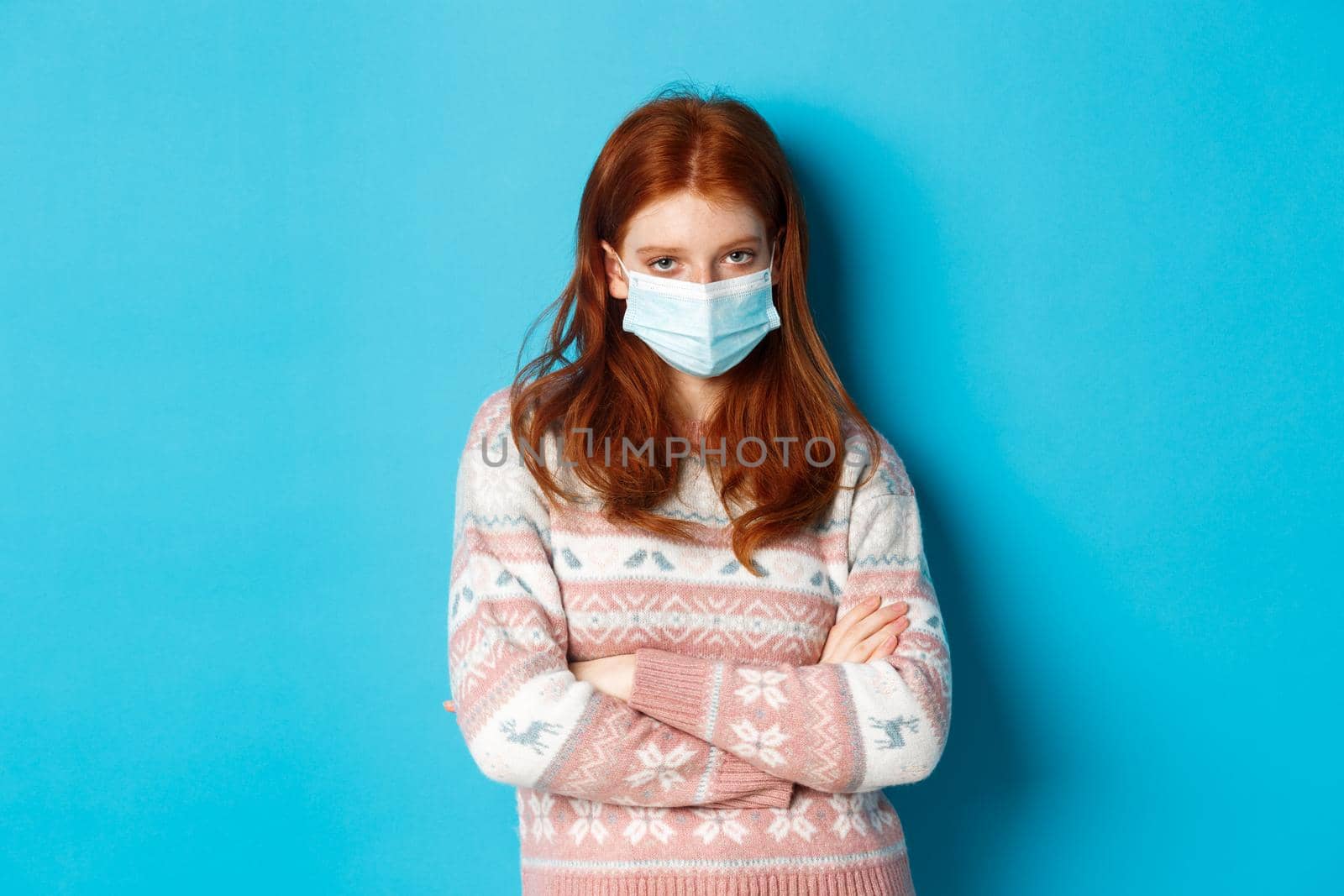 Winter, covid-19 and pandemic concept. Skeptical redhead girl in medical mask, cross arms on chest and stare angry at camera, standing over blue background.