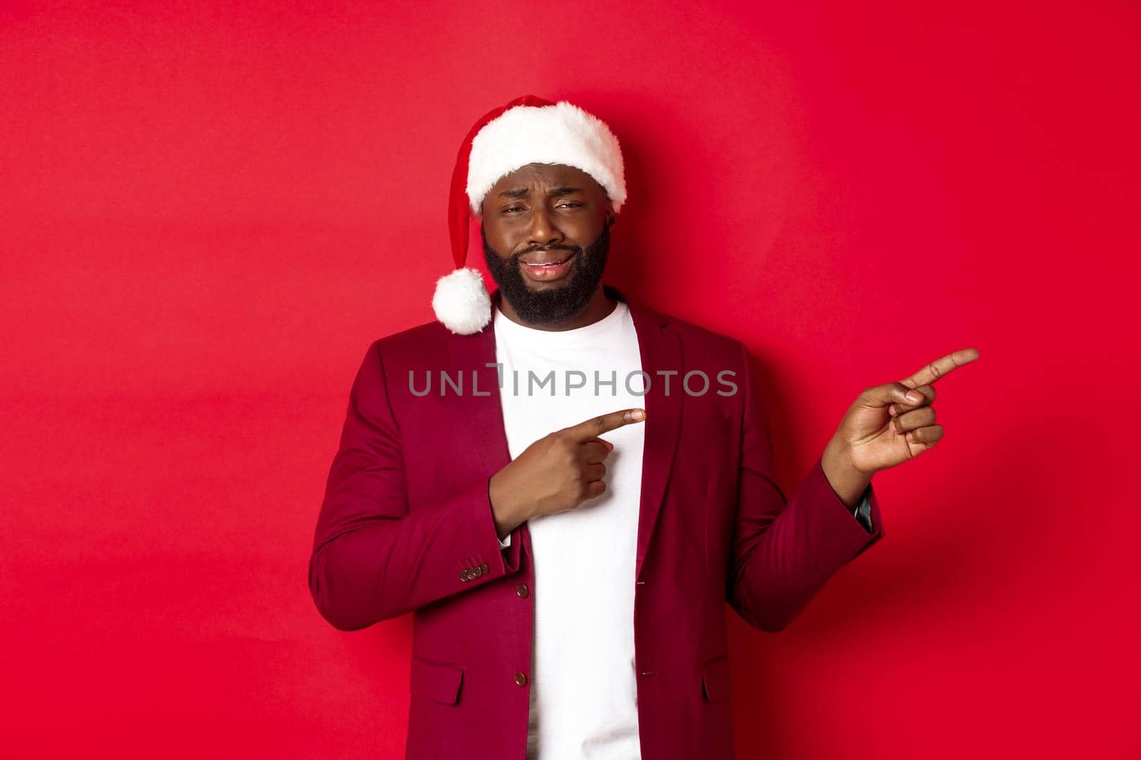Christmas, party and holidays concept. Skeptical and unamused Black man looking with disdain, pointing fingers right at logo, standing in santa hat against red background by Benzoix
