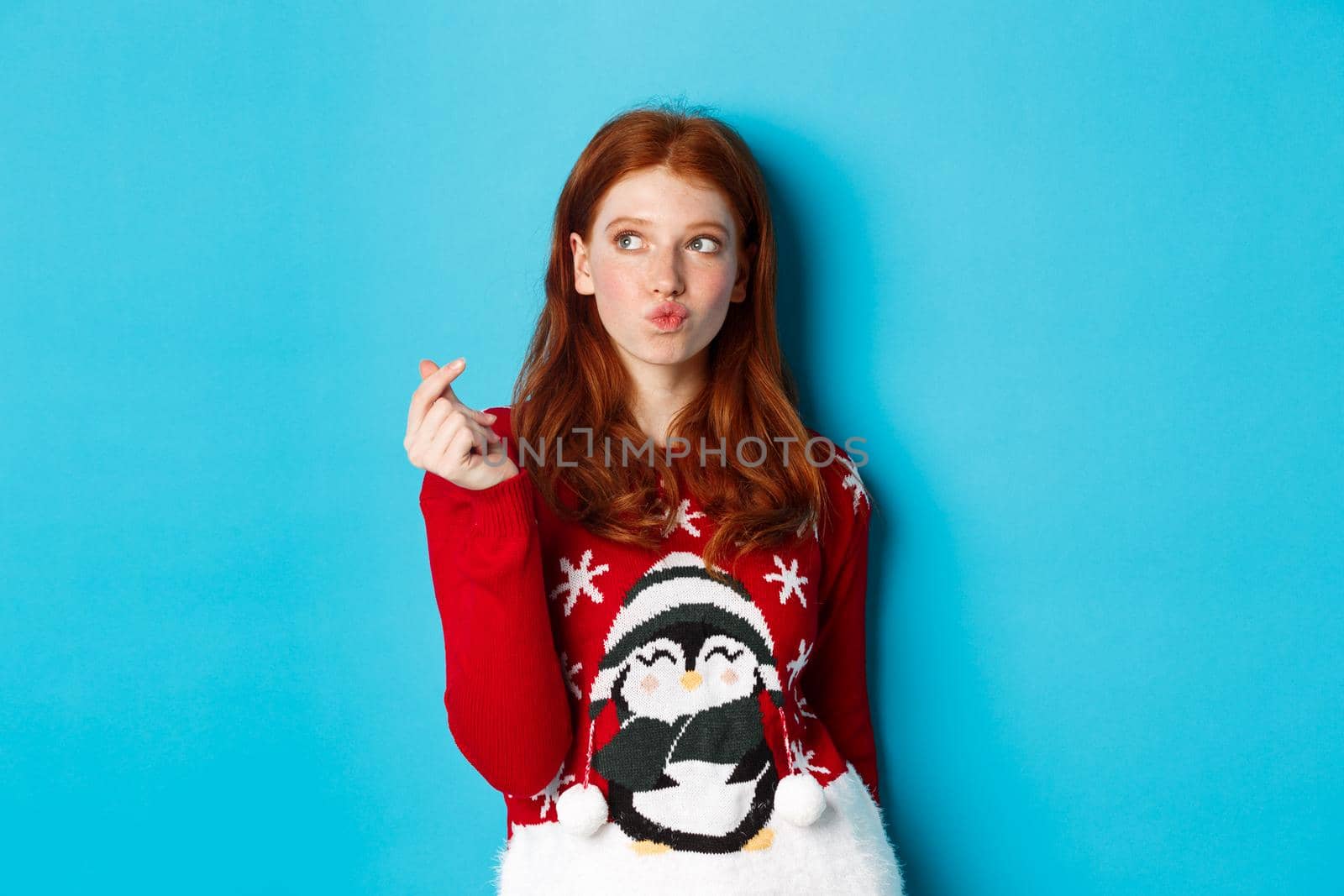 Winter holidays and Christmas Eve concept. Lovely redhead woman in xmas sweater, showing heart sign and thinking, looking upper left corner at logo, blue background.