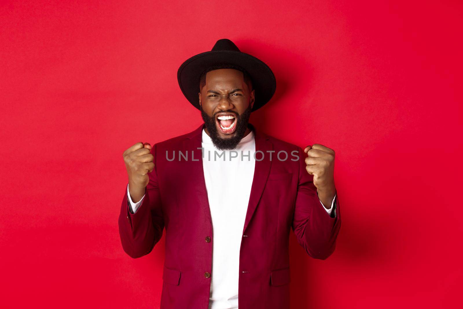 Satisfied lucky Black man celebrating victory, screaming from rejoice, winning prize, saying yes and fist pump, red background. Copy space