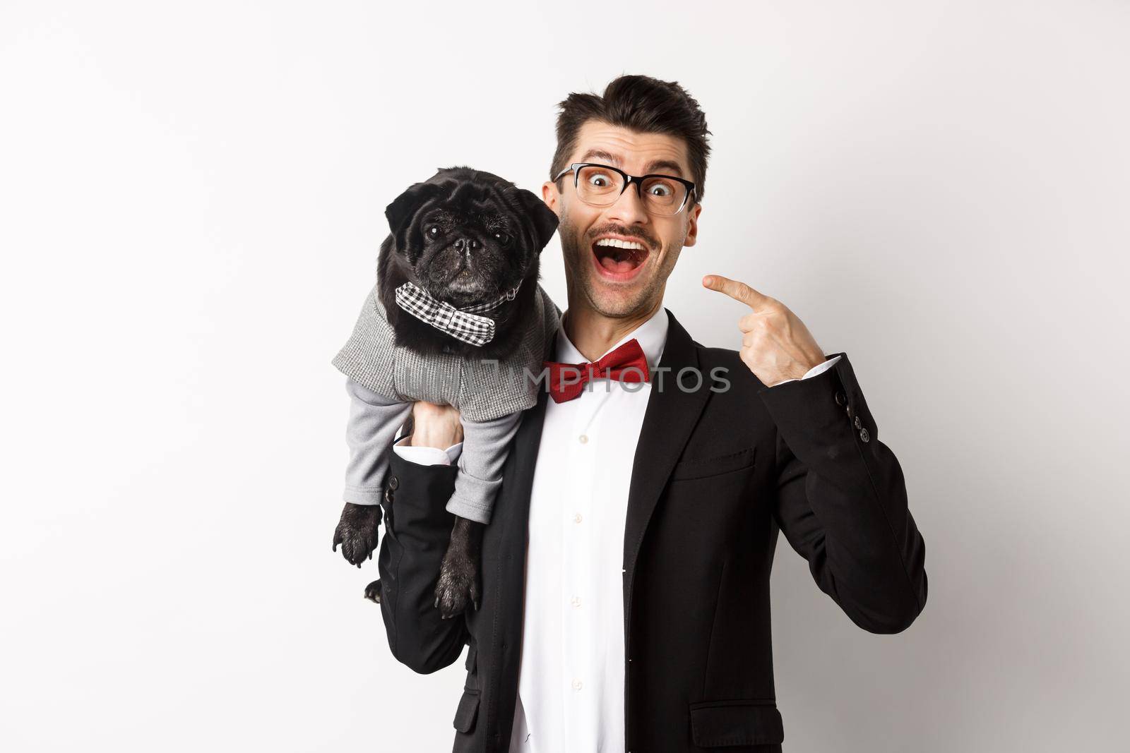 Amazed dog owner pointing at his cute black pug, smiling happy, puppy wearing costume, white background.