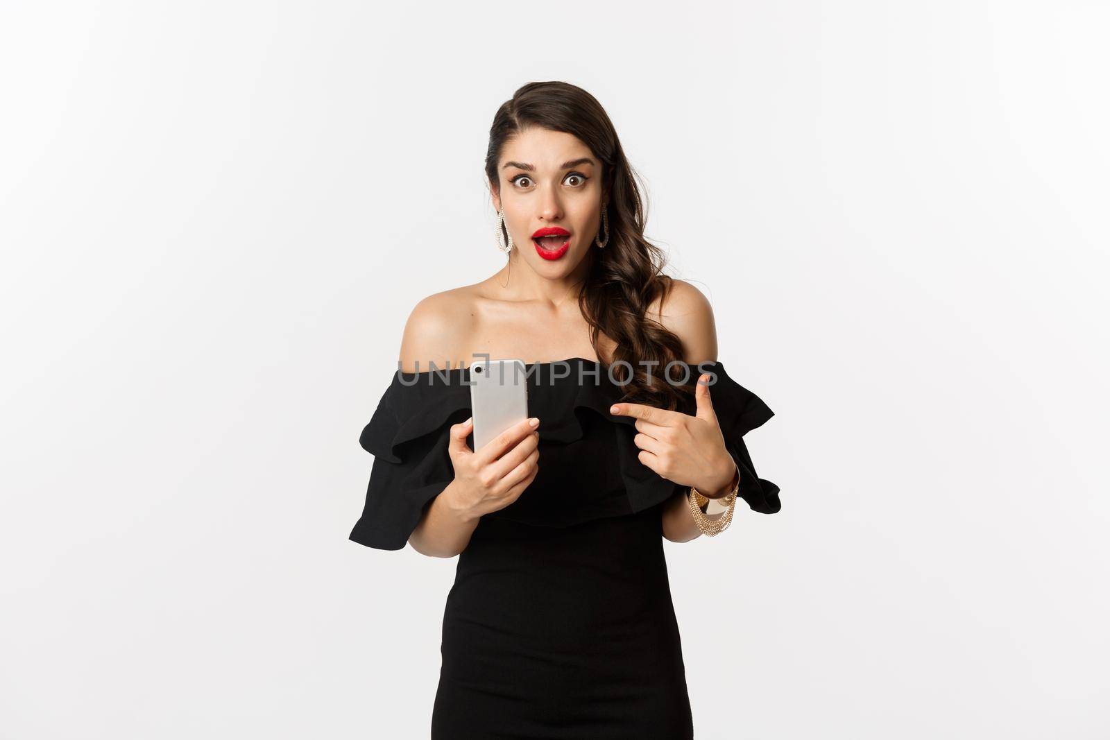 Online shopping concept. Stylish woman in black dress, wearing makeup, pointing finger at mobile phone with surprised emotion, standing over white background.
